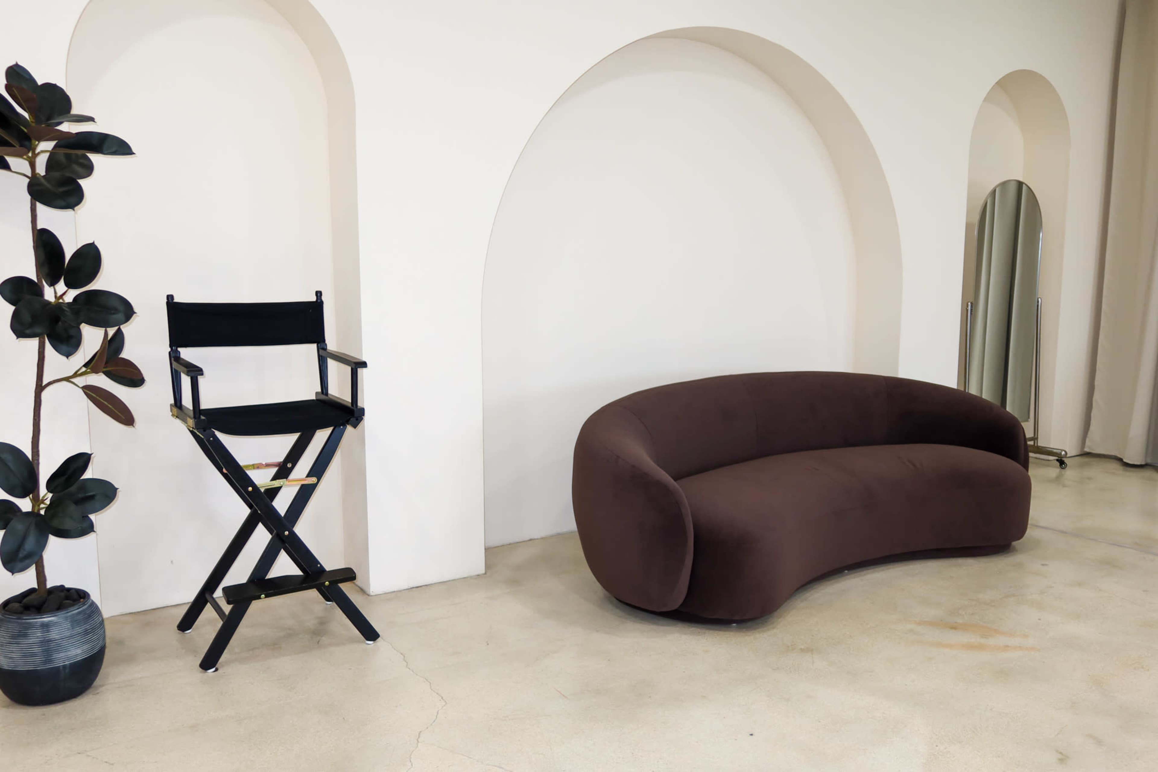The scene features a curved brown sofa next to a black director's chair, with a potted plant and a full-length mirror against a backdrop of arched wall segments.