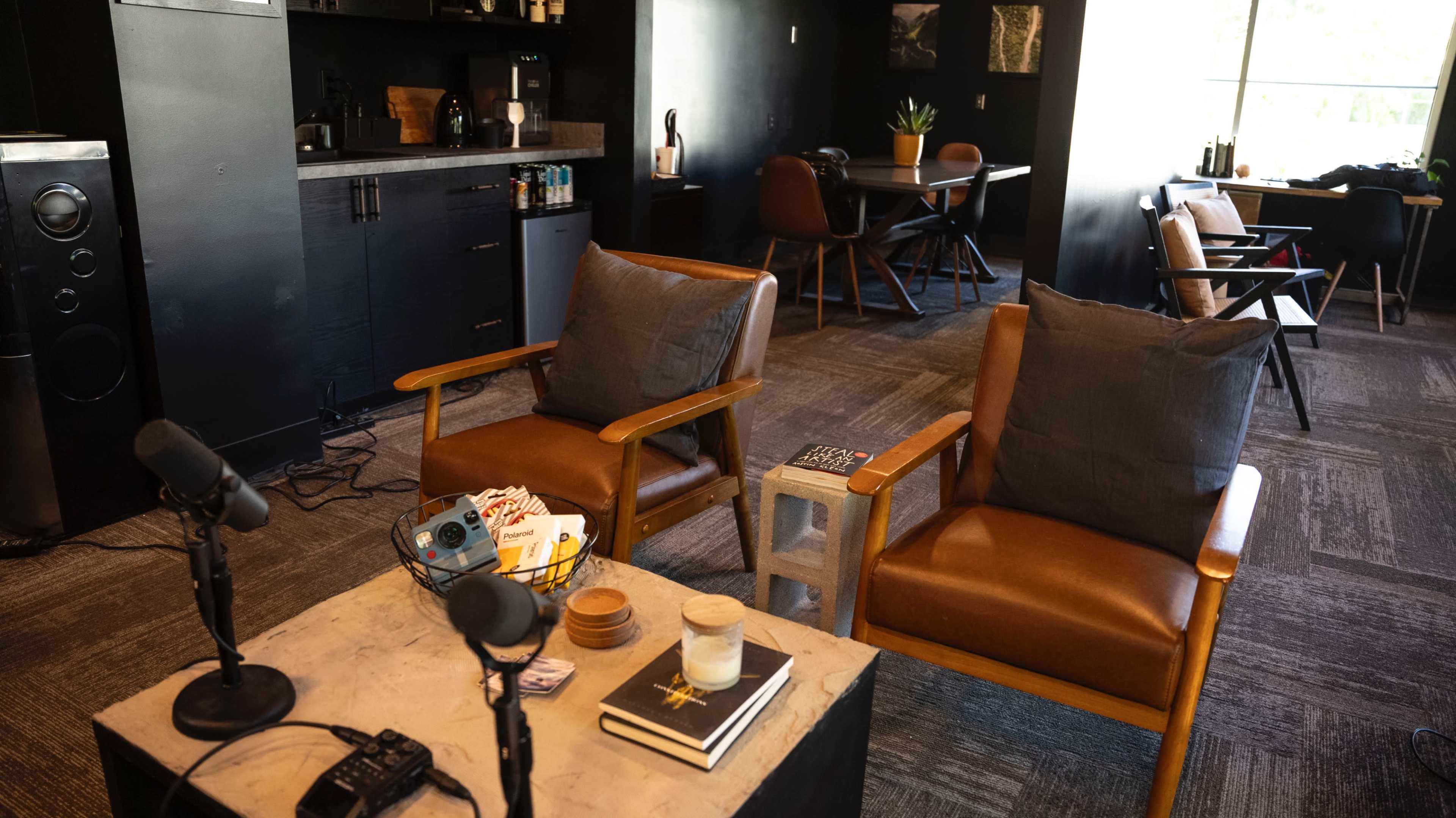 The image shows a cozy interior space with two brown chairs, a coffee table with books and decor, and a small kitchenette area in the background.