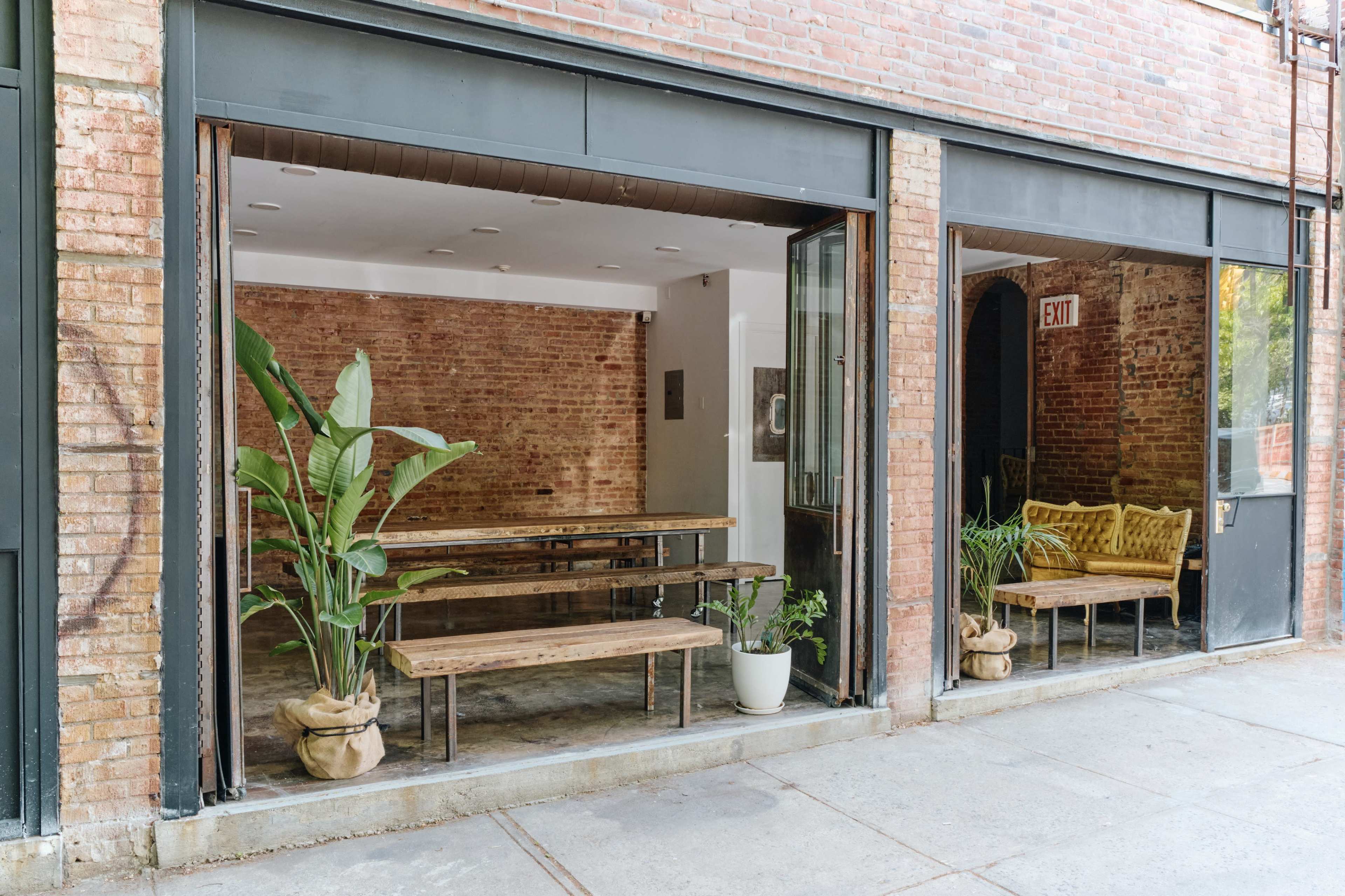 A modern, open storefront with large glass windows revealing an interior featuring brick walls, wooden benches, and potted plants.