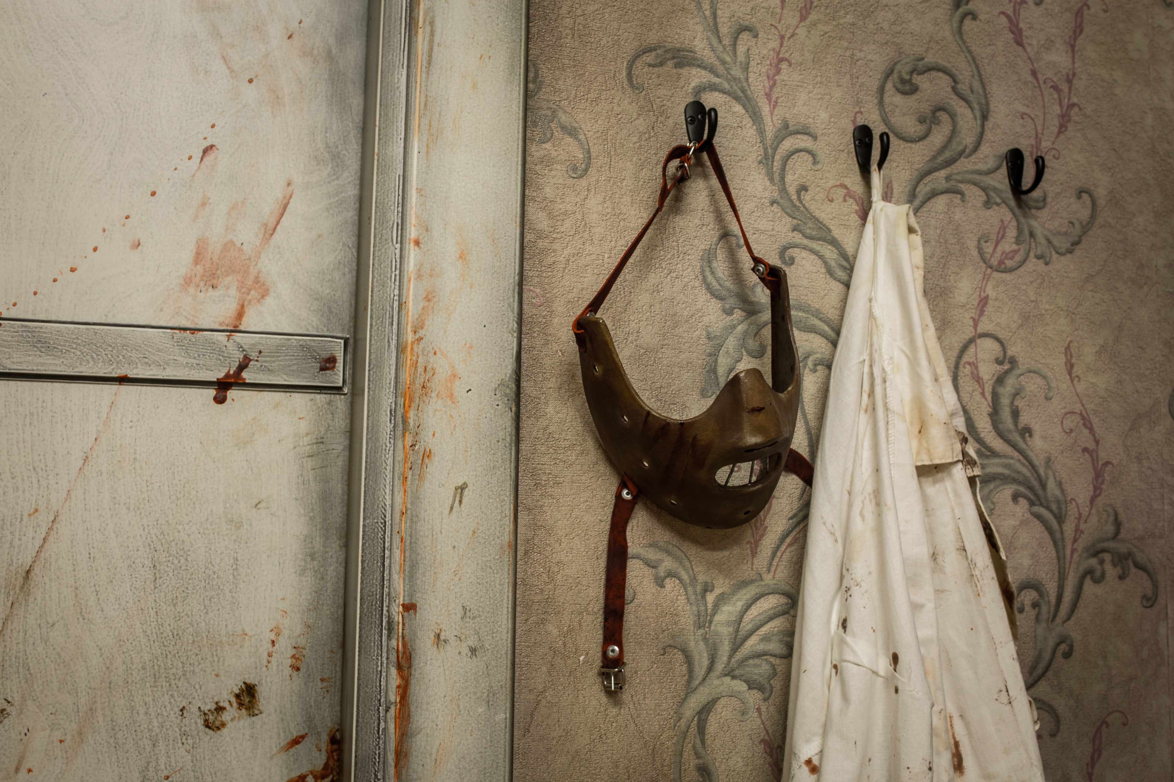 A weathered mask hangs on a wall beside a soiled white coat, set against a patterned backdrop with stains.