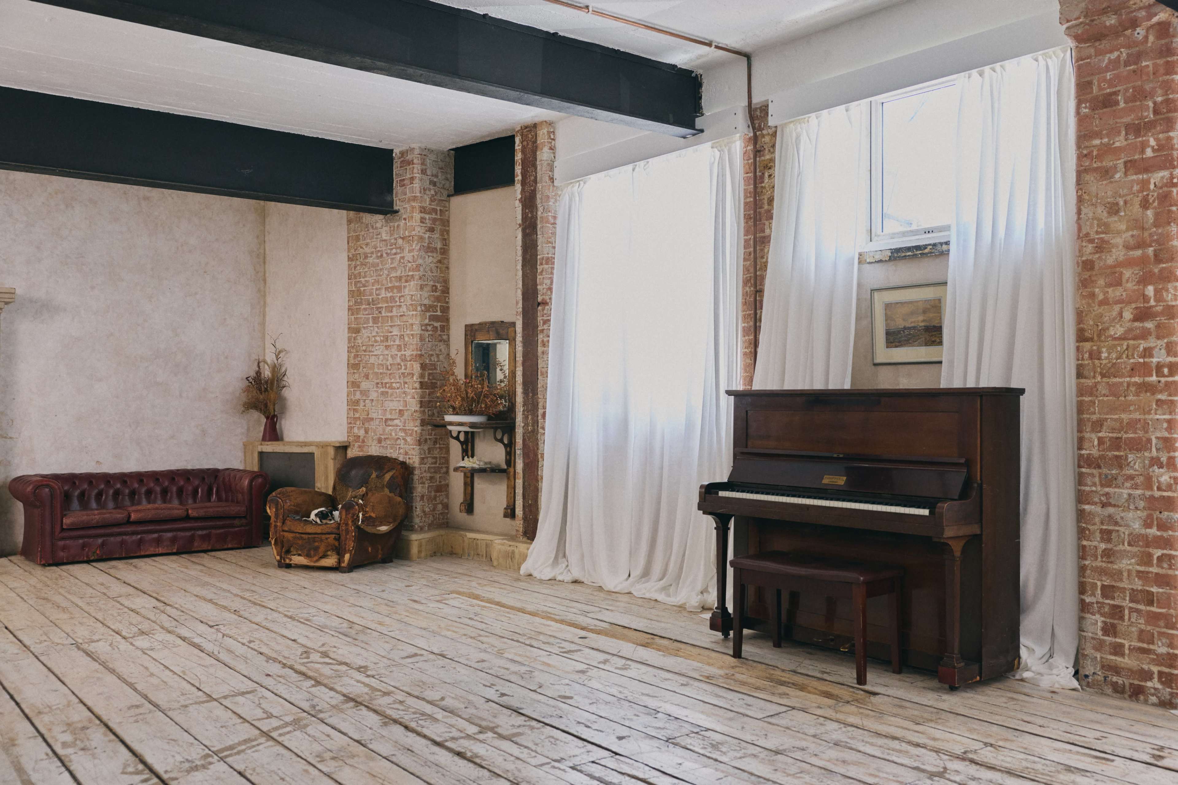 The image shows a spacious room with exposed brick walls, a wooden floor, a vintage piano, a dark sofa, and floor-to-ceiling curtains beside a window.