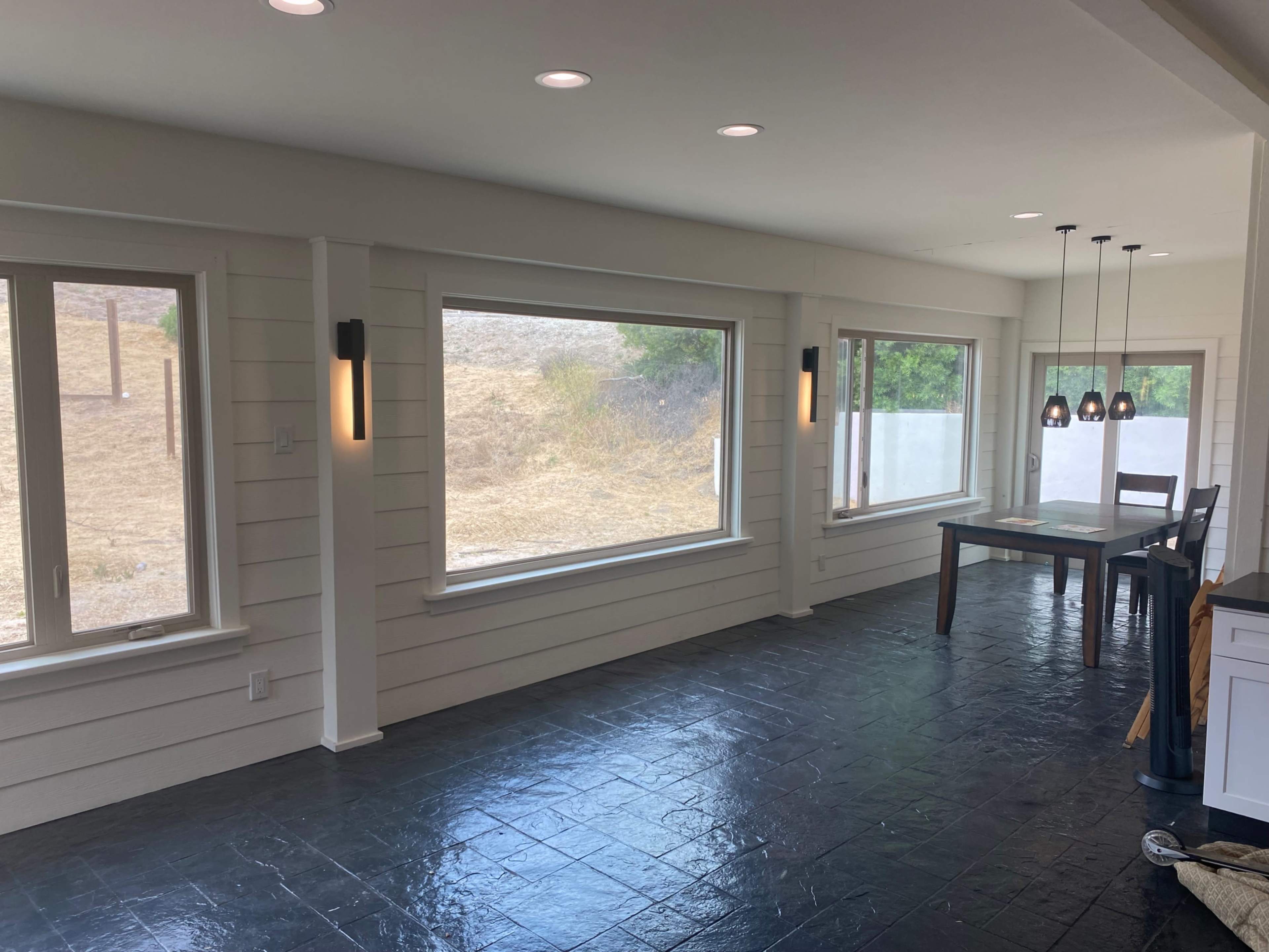 The image shows a spacious interior room featuring large windows along one wall, a dining table and chairs in the corner, and slate flooring.