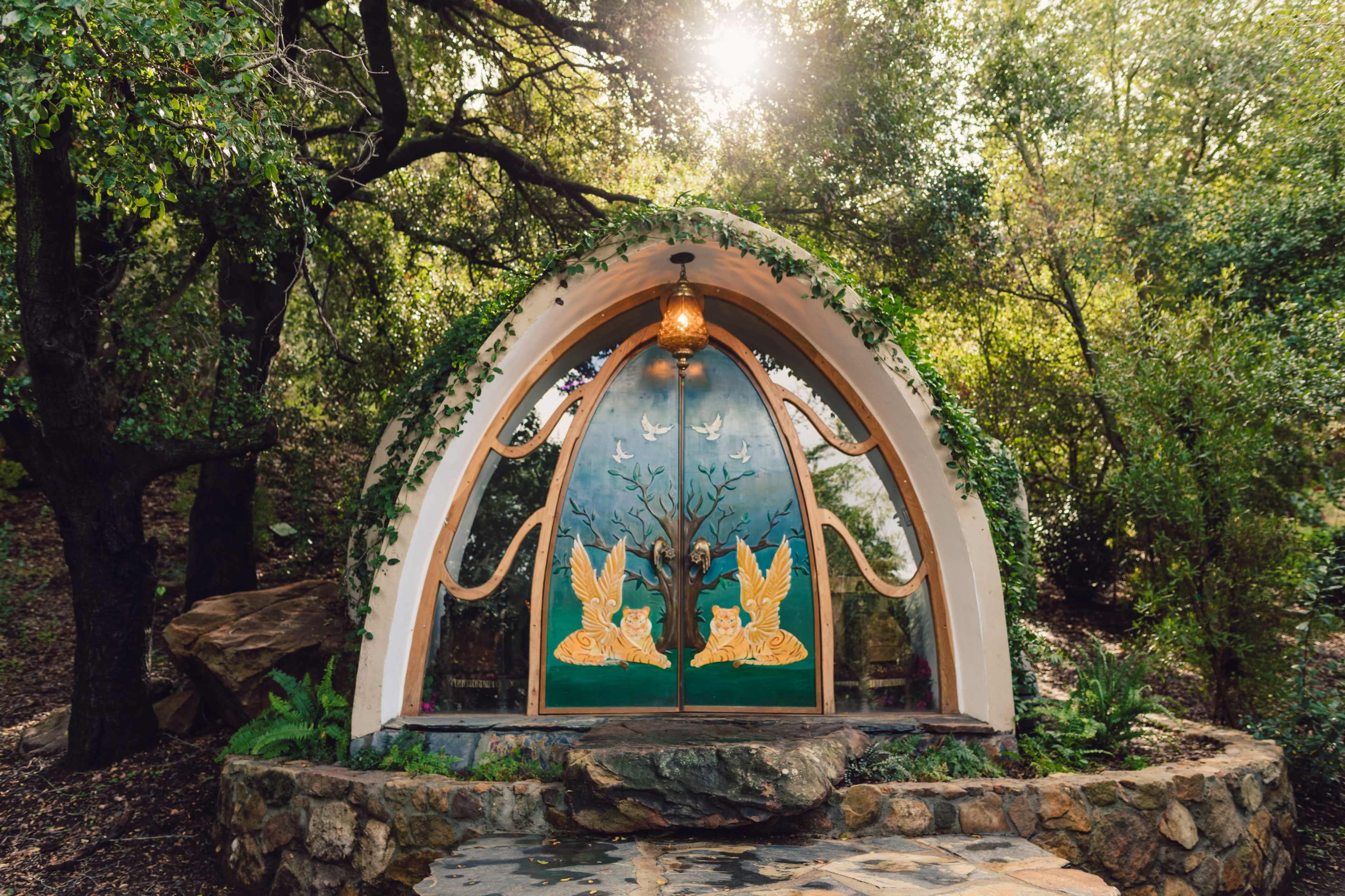 A decorative entrance features ornate wooden doors with bird carvings, surrounded by lush greenery and sunlight filtering through the trees.