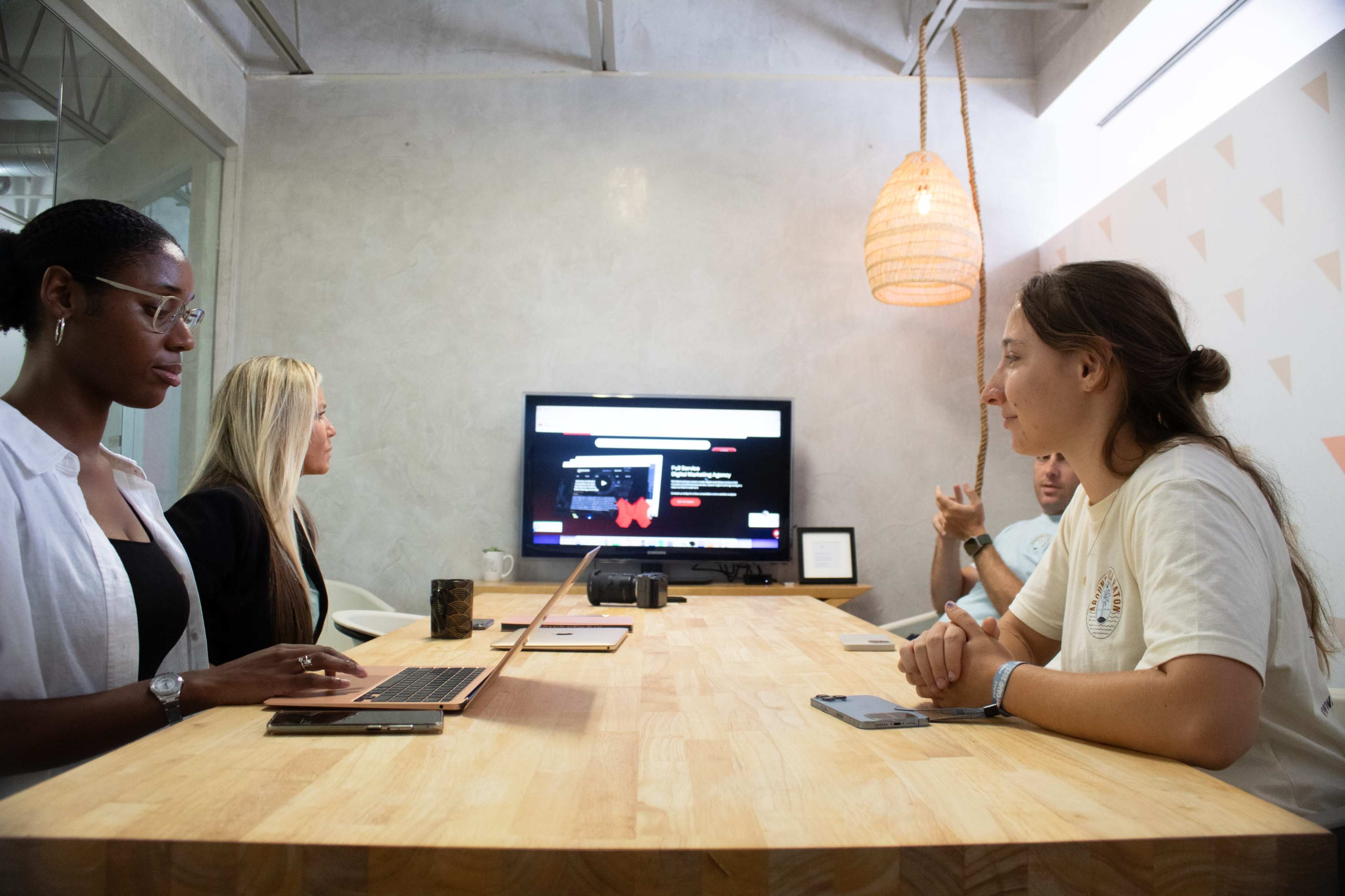 A group of four people holds a meeting around a wooden table in a modern conference room with a screen displaying content in the background.
