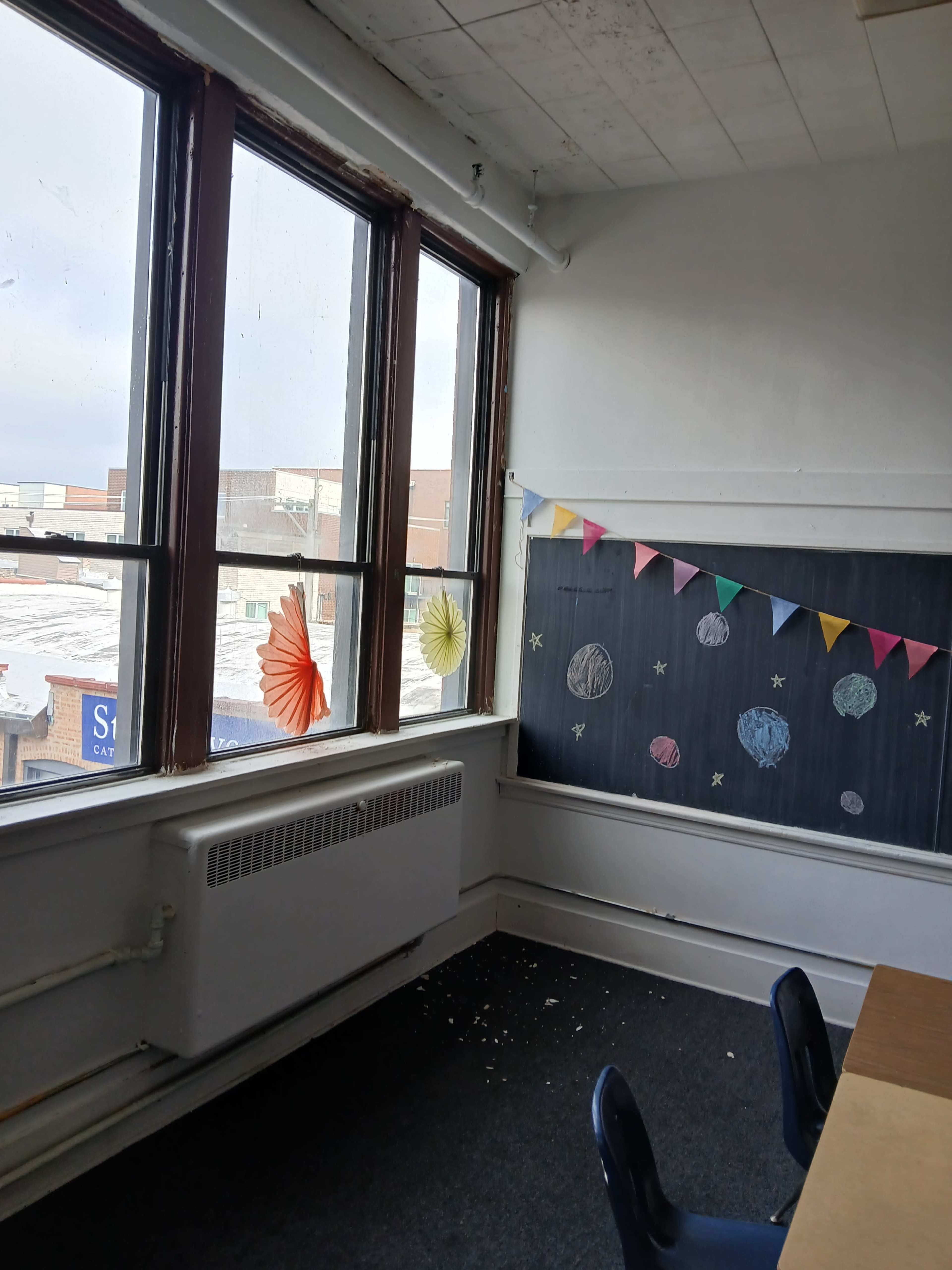 The image shows a classroom corner with large windows, a chalkboard decorated with planets and colorful bunting, and a carpeted floor.