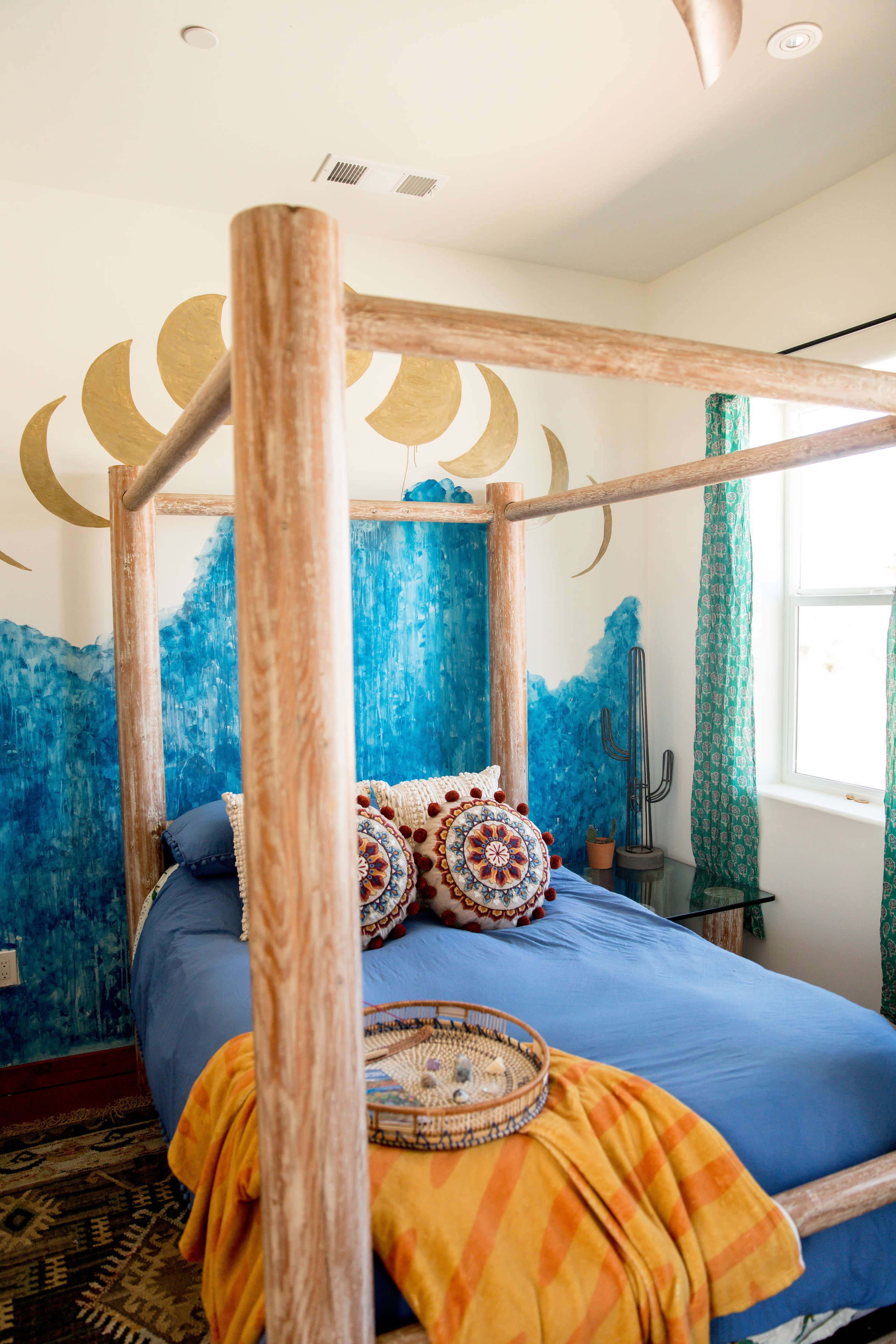 A four-poster bed with a blue comforter is positioned against a wall decorated with a blue and gold mural, featuring decorative pillows and a tray on the bed.