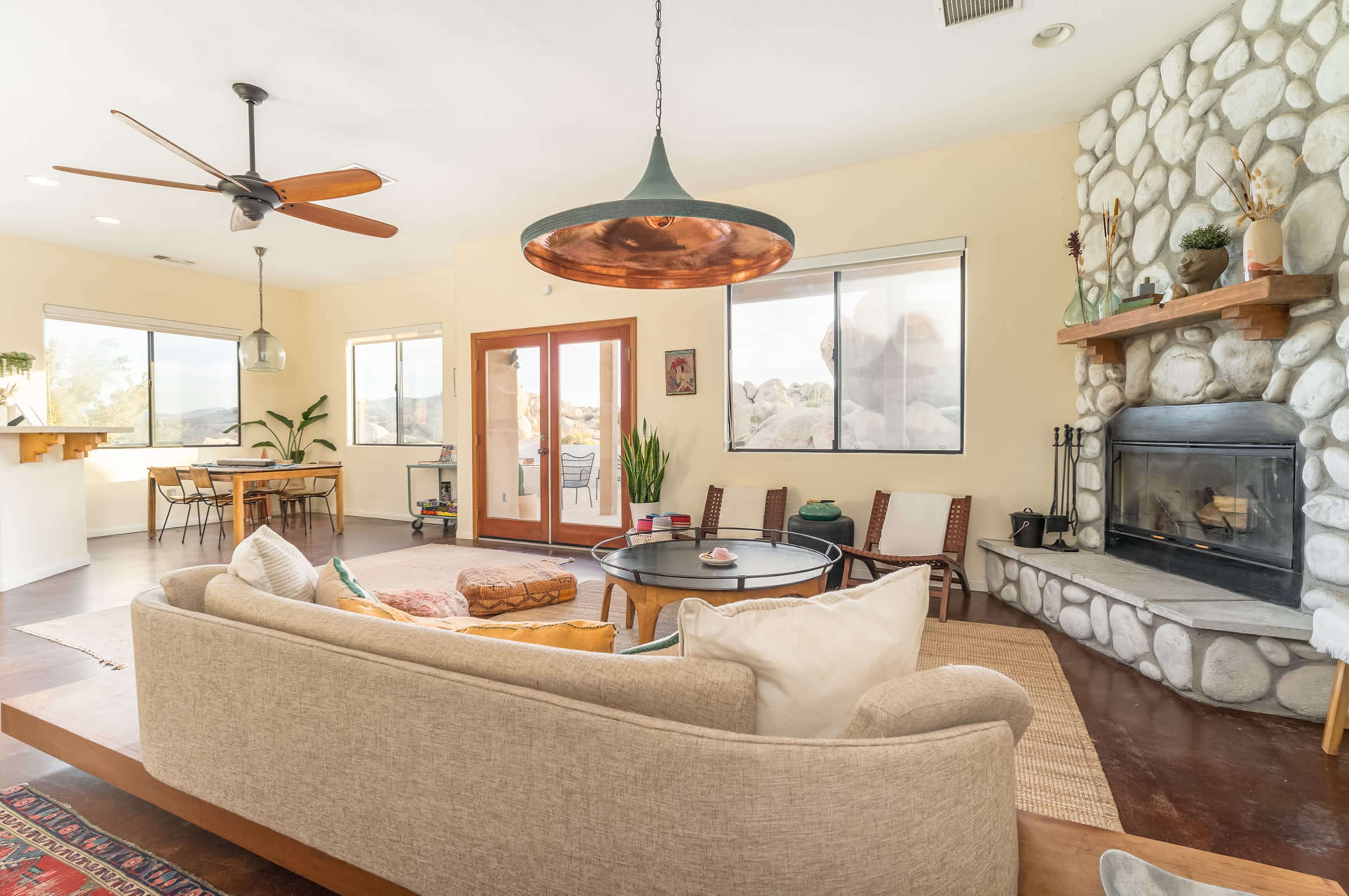 A modern living room with a stone fireplace, large windows, and a dining area featuring a wooden table and chairs.