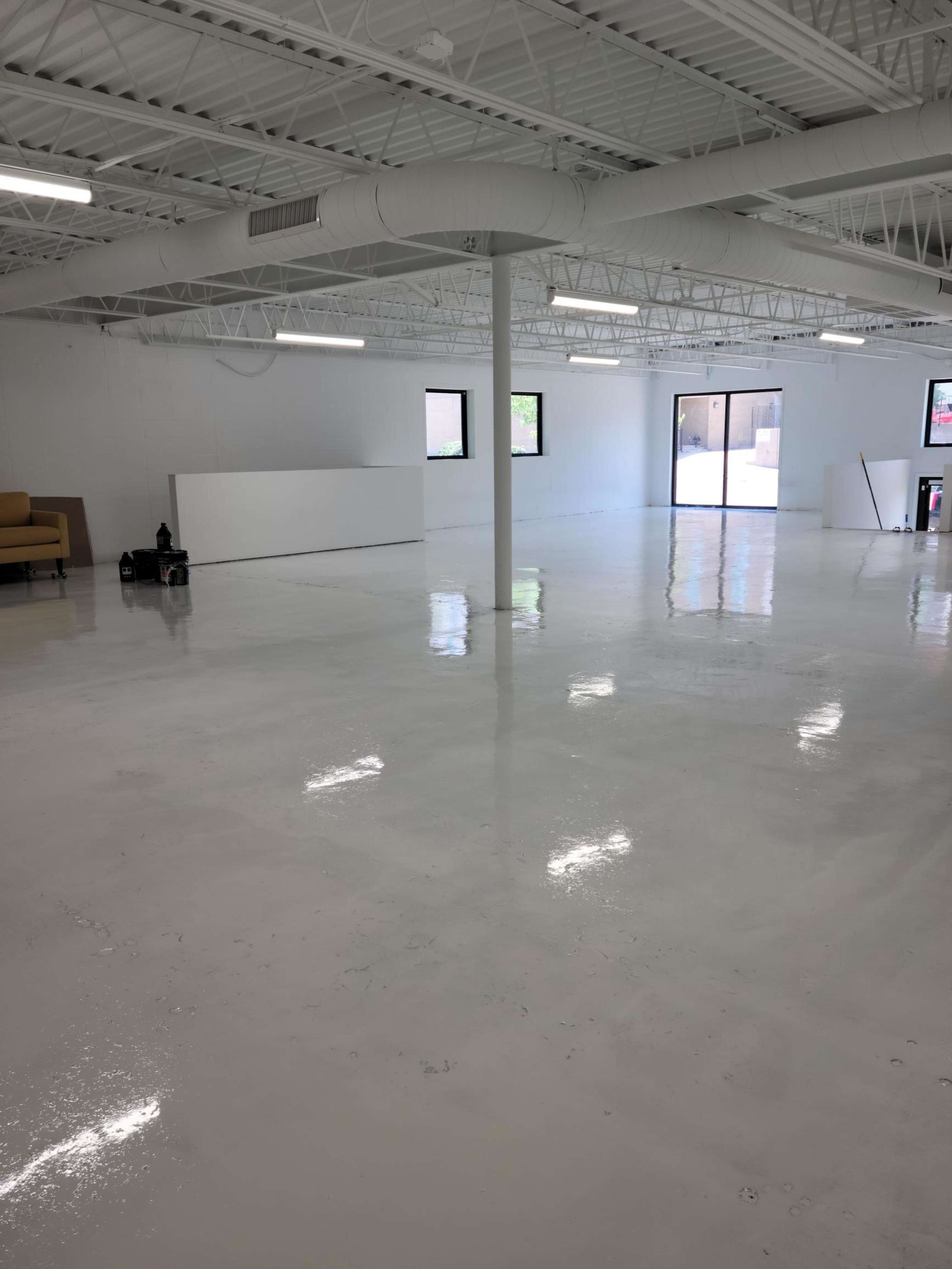 A spacious, empty room with a glossy white floor, white walls, and large windows, featuring minimal furnishings.