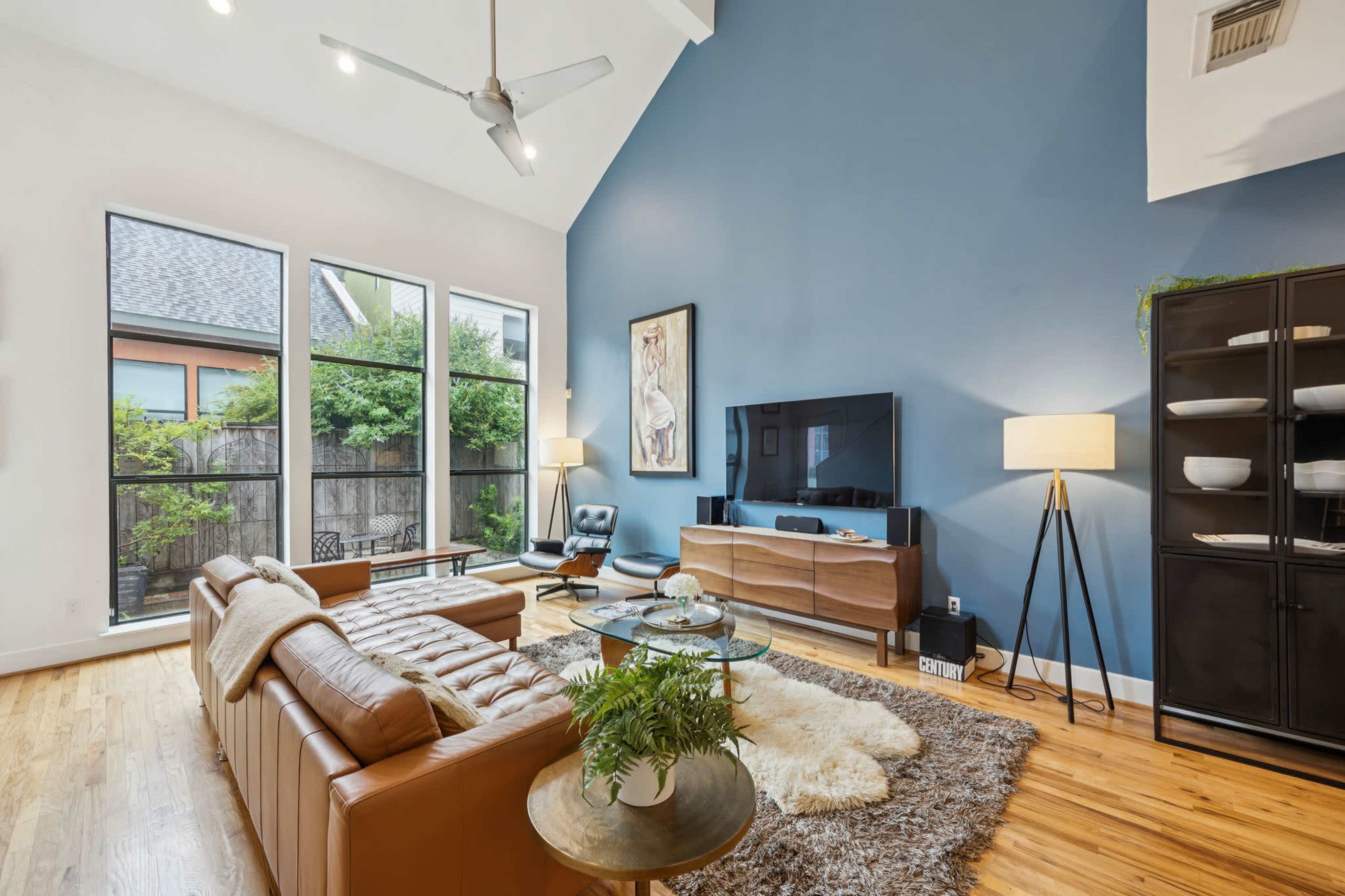 The image shows a modern living room with a high ceiling, featuring a brown leather sofa, a glass coffee table, and a large television against a blue wall, with large windows allowing natural light to enter.