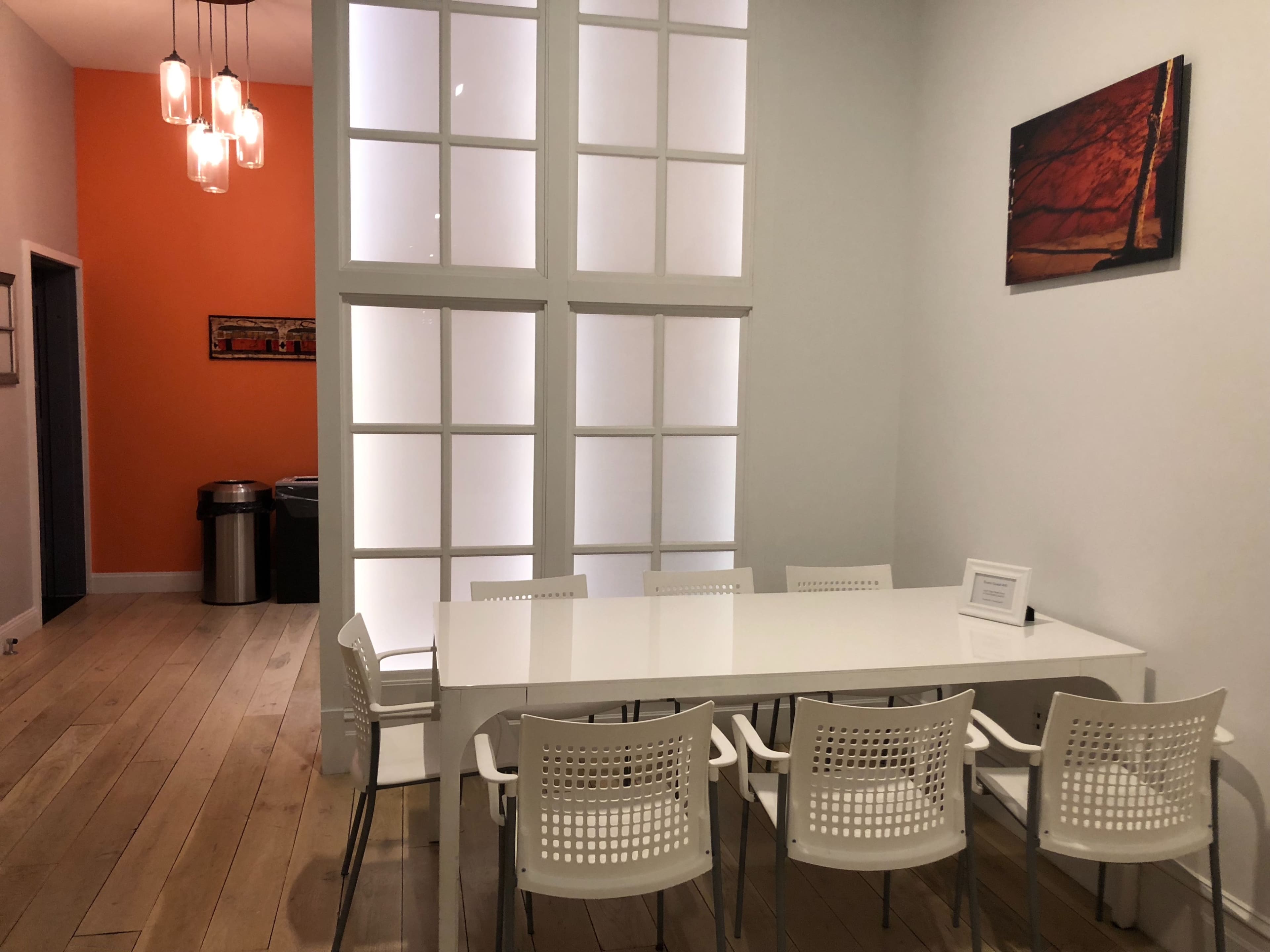 A modern conference room with a white table surrounded by six chairs, framed by a wall of glass panels and an orange accent wall.