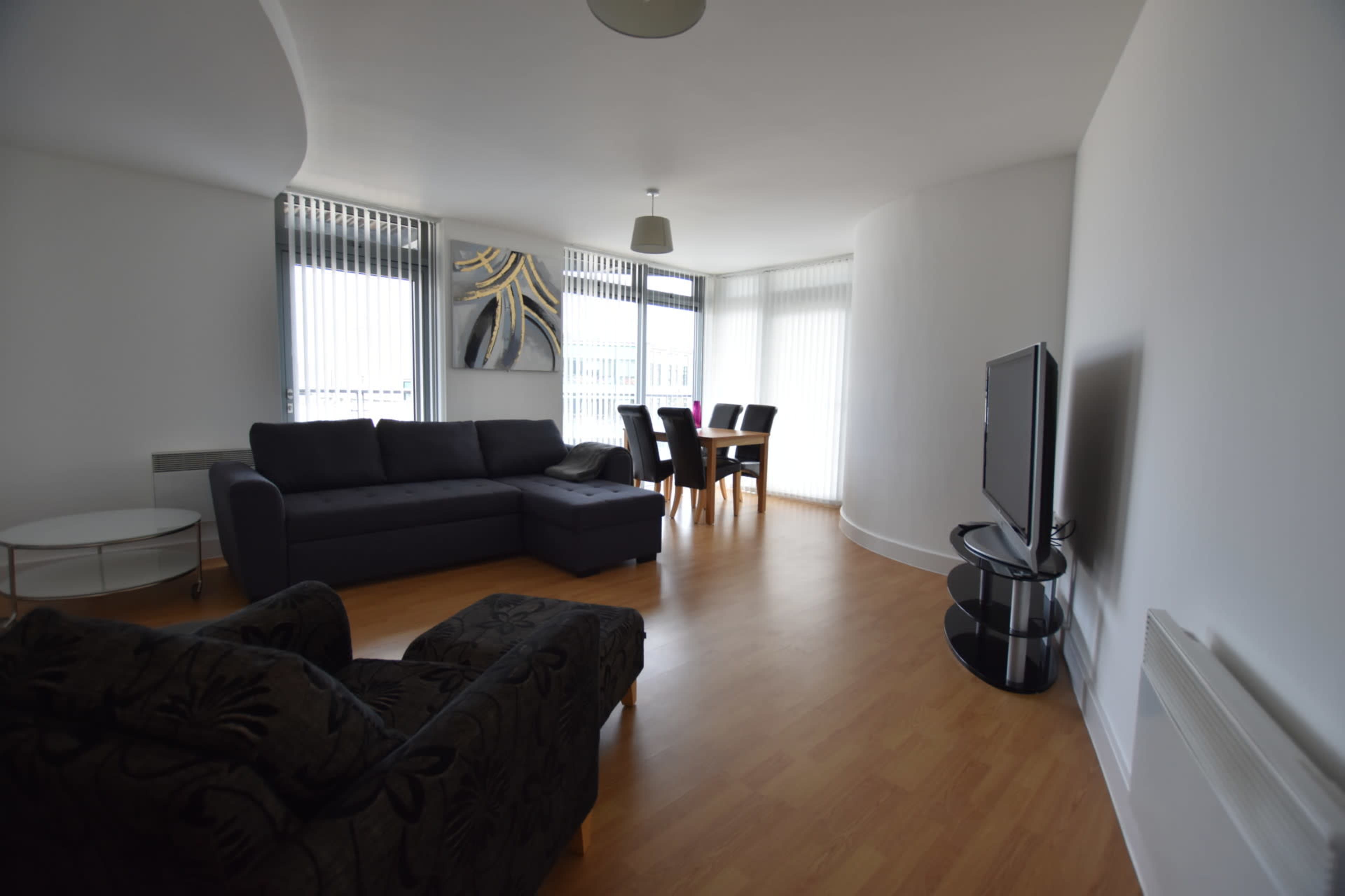 A modern living room with a dark sofa, a black armchair, a dining table with chairs, and a television on a stand, all set against a bright, minimalist interior.