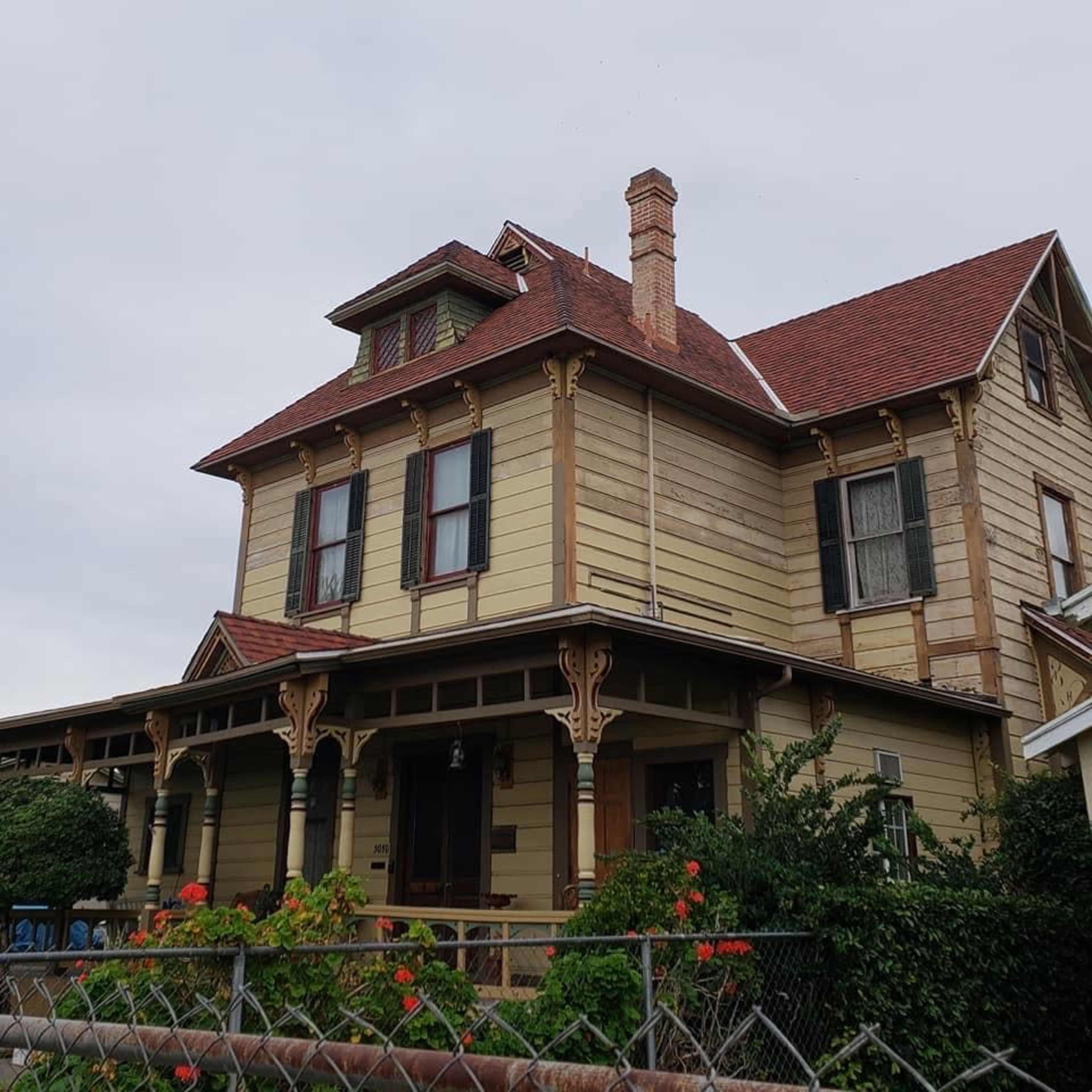 A two-story Victorian-style house with a gabled roof, ornate trim, and a front porch surrounded by a fence and garden.