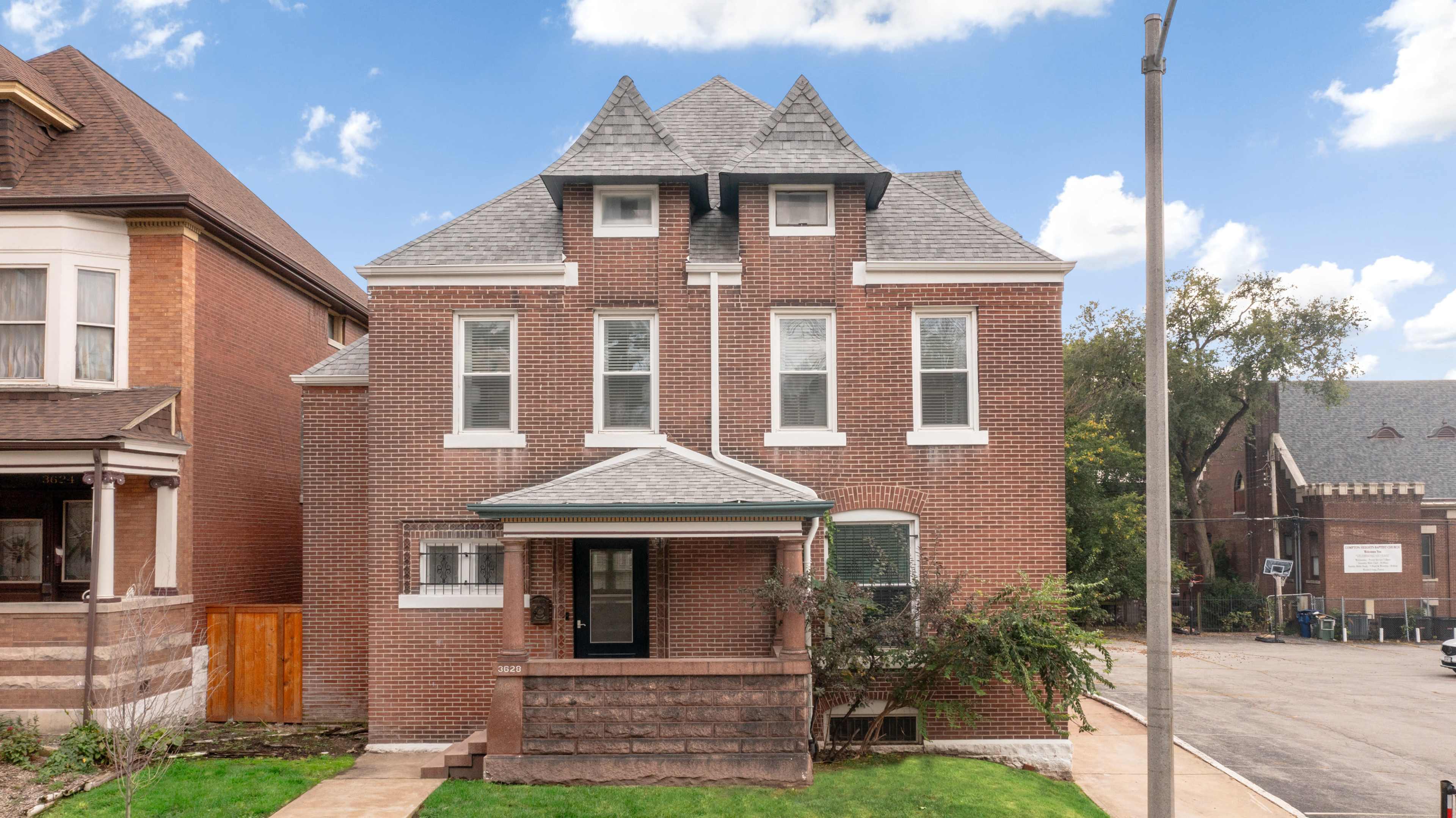 A two-story brick house with a distinctive gabled roof and a porch, situated on a residential street.