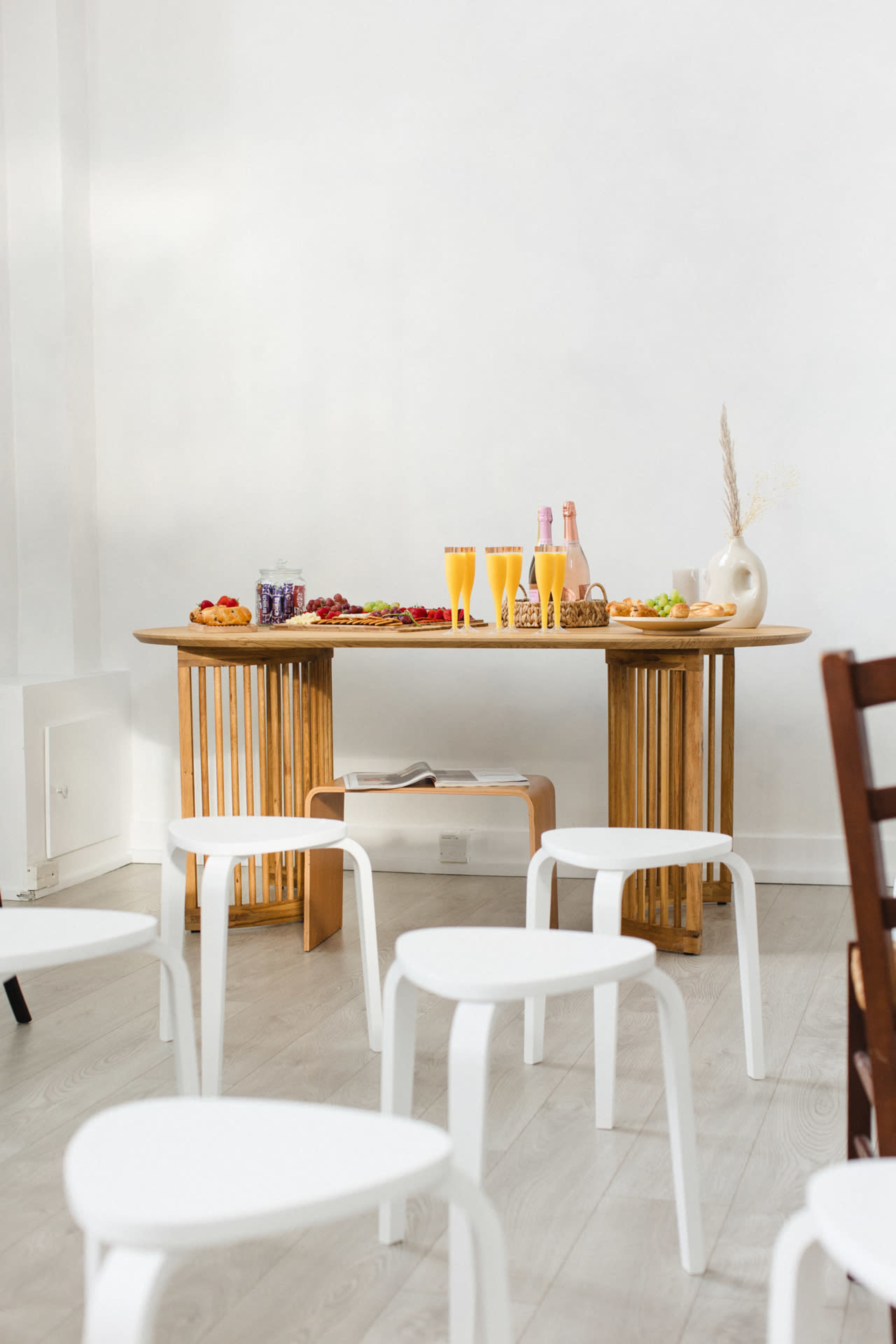 A wooden table is set with a variety of food and drinks, including glasses of orange juice and sparkling beverages, surrounded by white chairs.