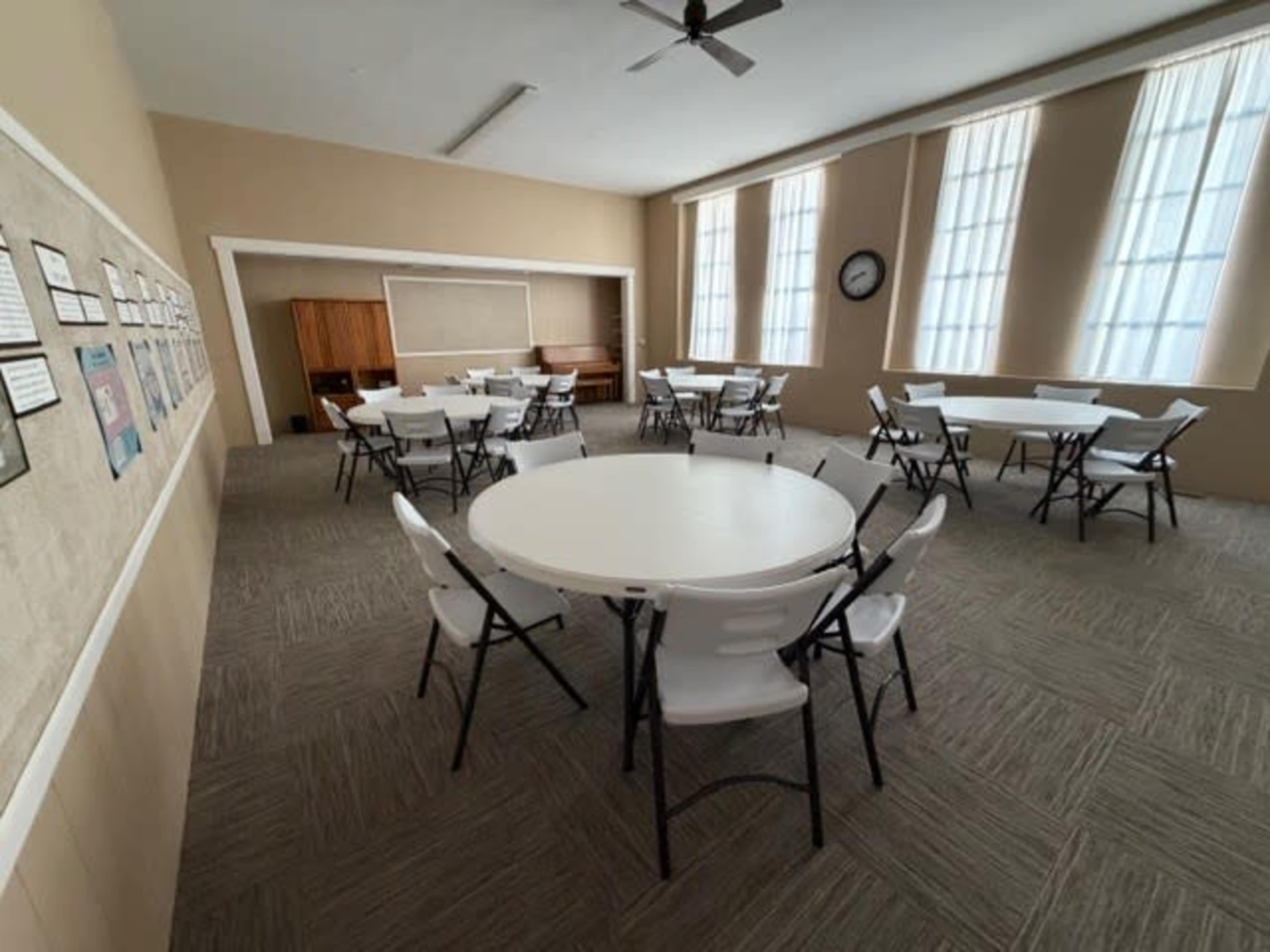 The image shows a quiet, spacious room with multiple round tables and chairs arranged neatly, featuring large windows and a wall displaying framed documents.