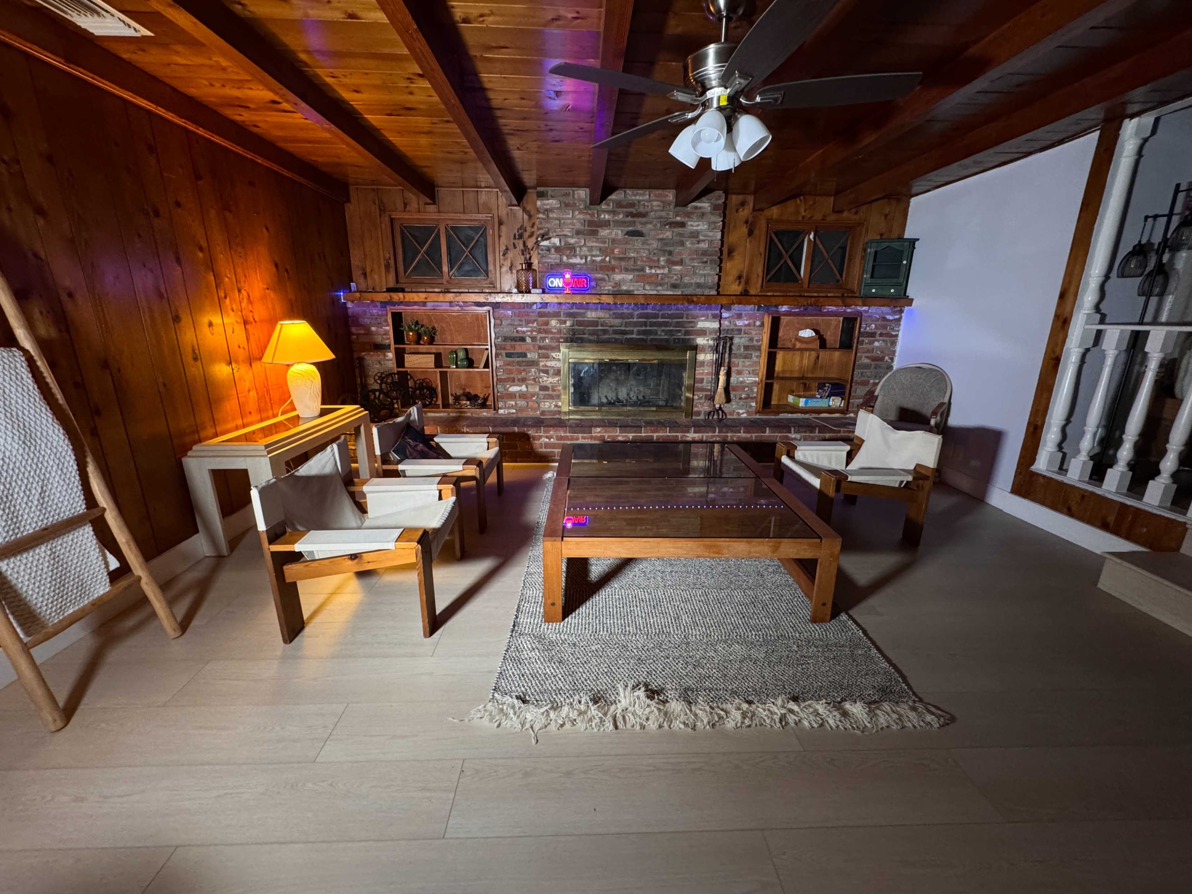 The image shows a cozy living room with wooden walls, a brick fireplace, and four wooden chairs arranged around a glass coffee table on a rug.