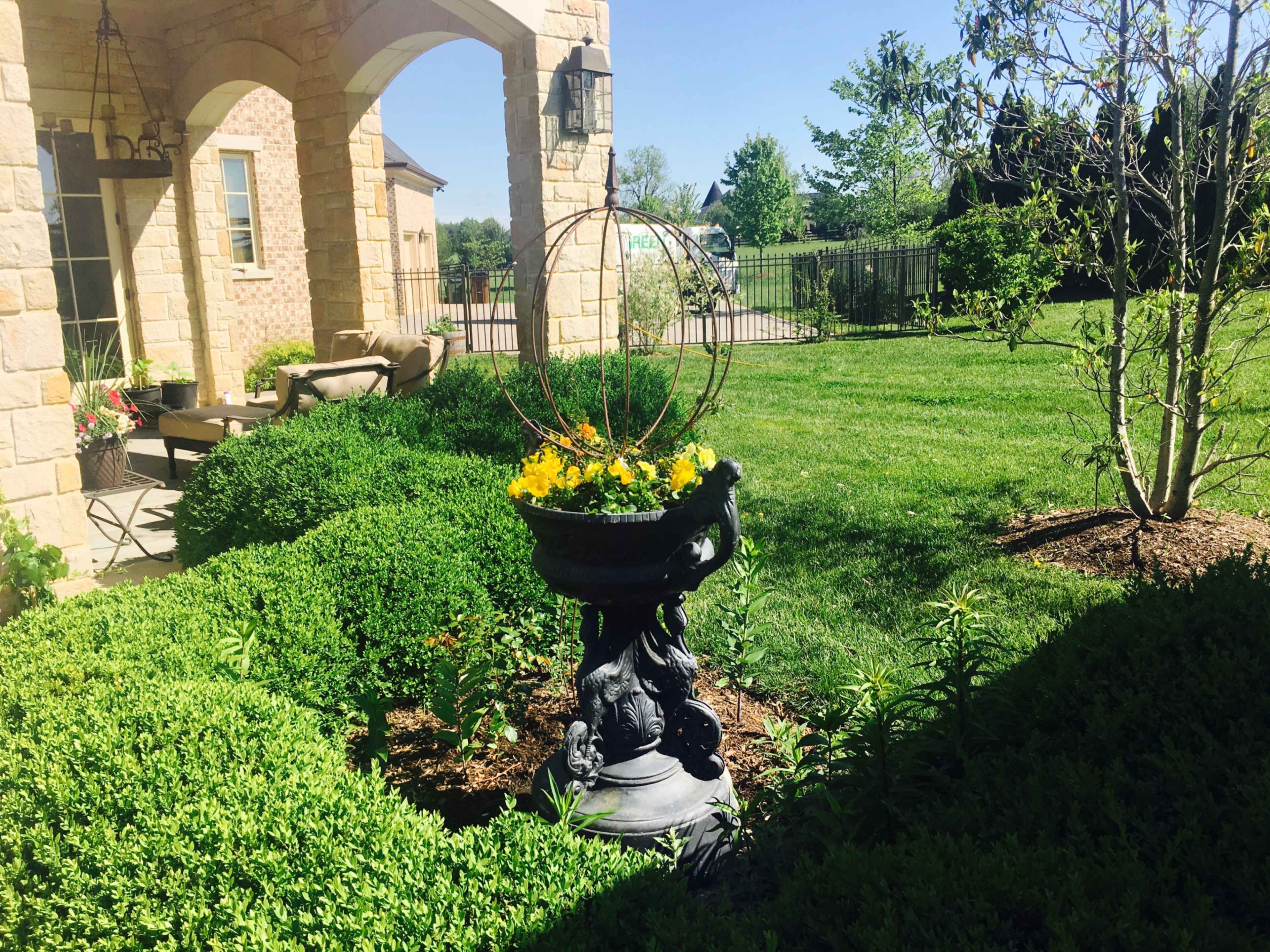 A stone planter with yellow flowers sits on a pedestal in a well-manicured lawn surrounded by hedges.
