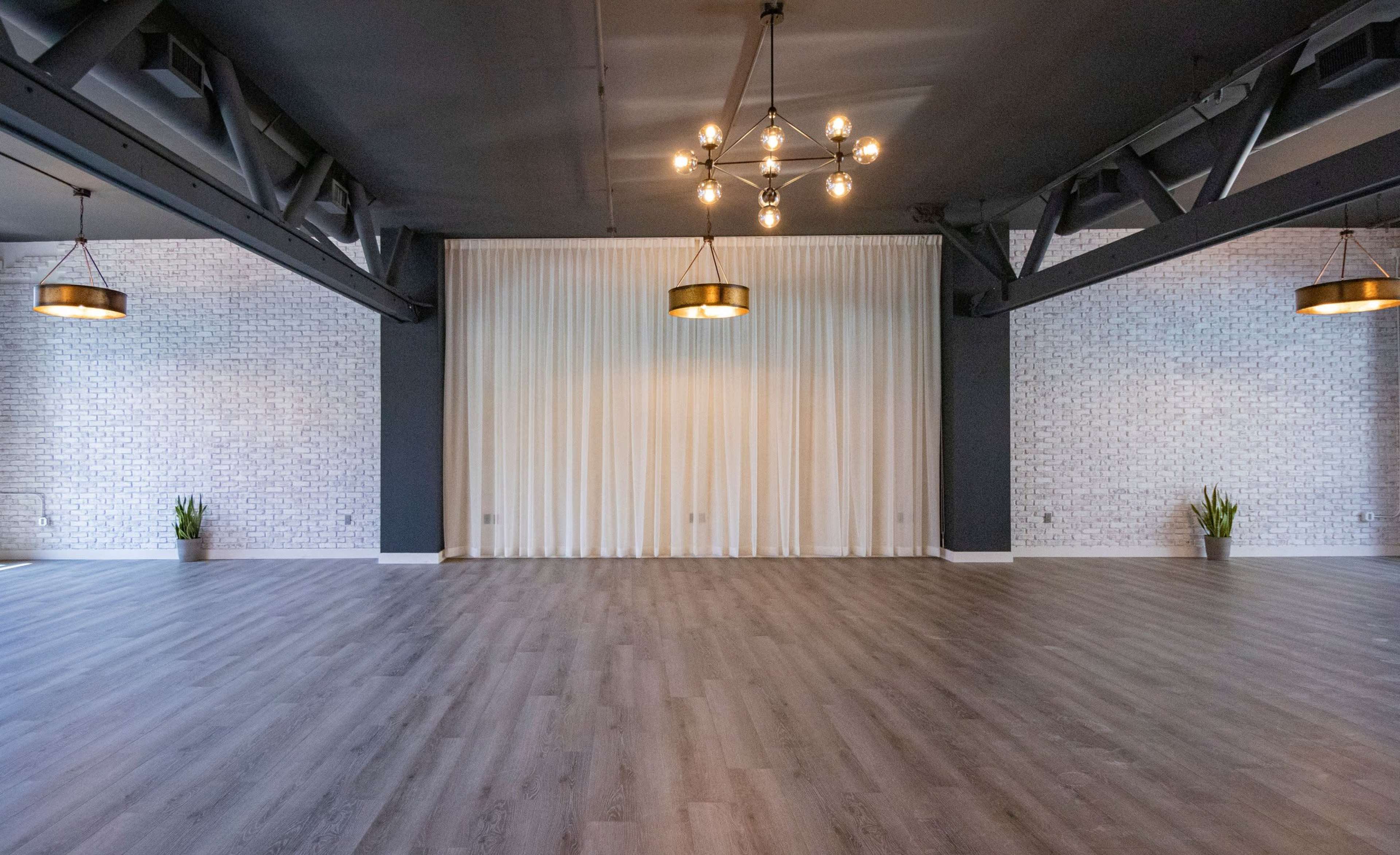 An empty hall features a wooden floor, a backdrop of white curtains, and industrial-style lighting fixtures.