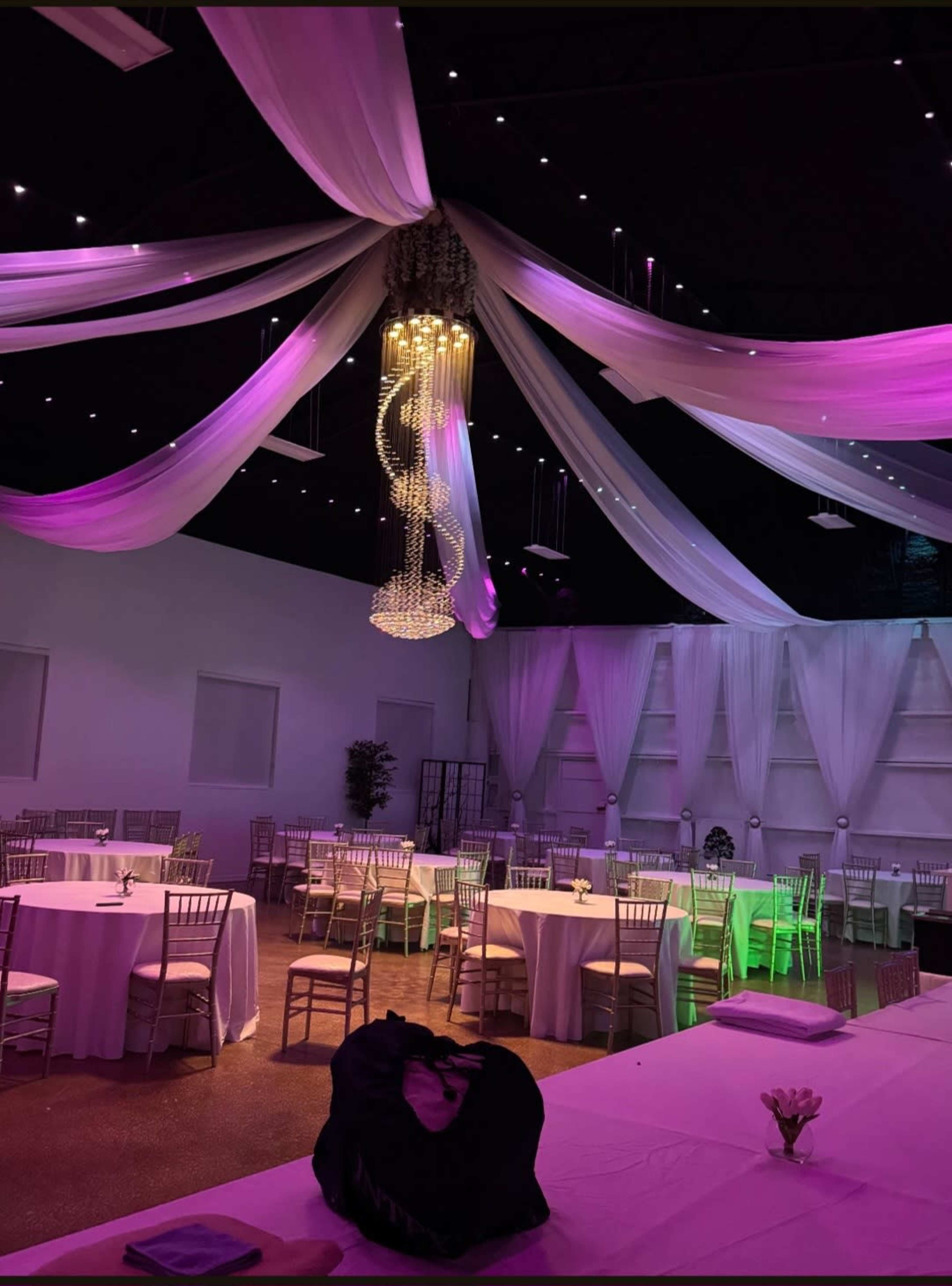 The image shows an elegantly decorated event space with round tables covered in white tablecloths, adorned with chairs, and draped fabric hanging from the ceiling, illuminated by soft purple lighting.