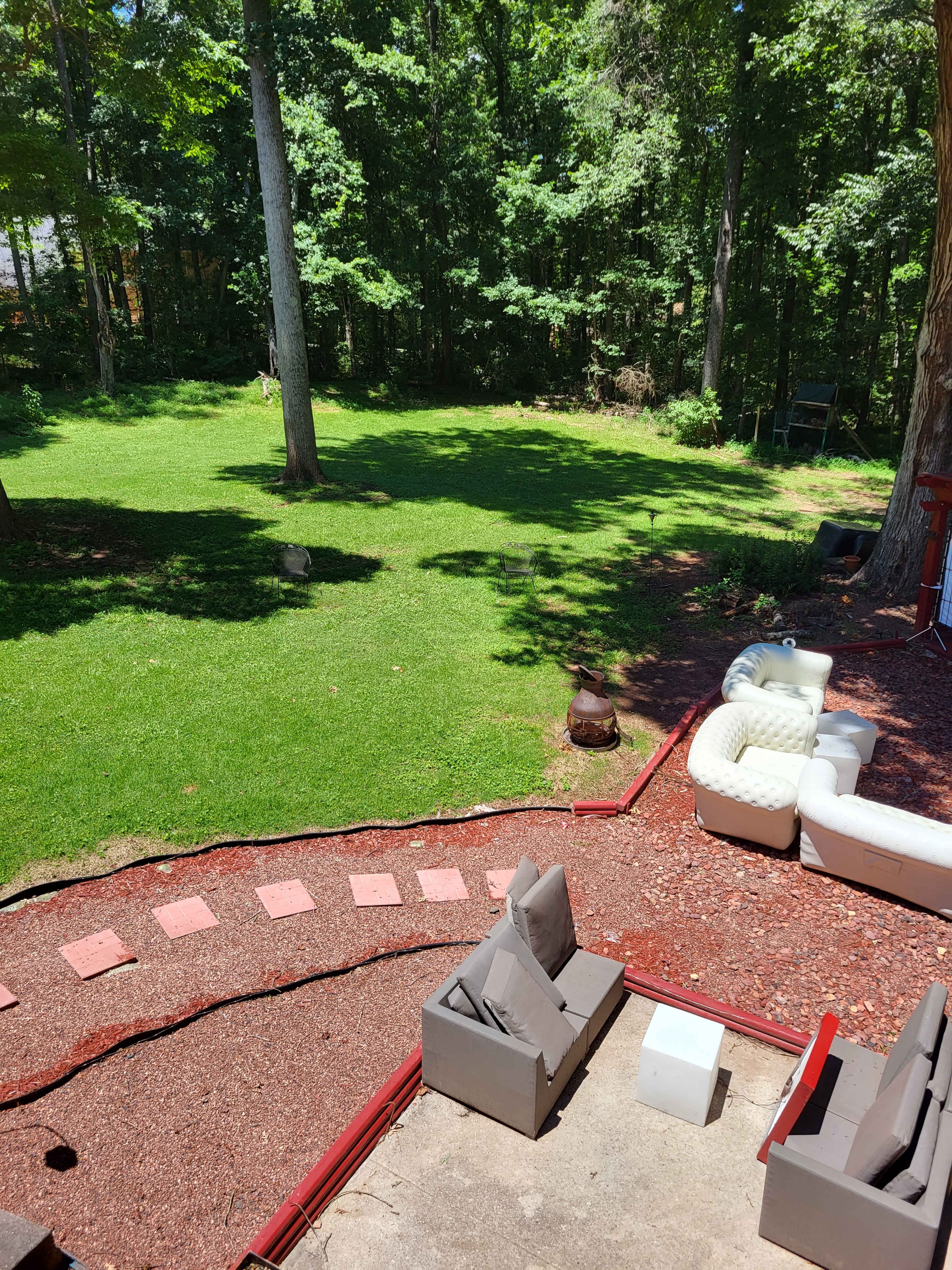 The image shows a backyard with green grass, a stone pathway, outdoor seating, and trees in the background.
