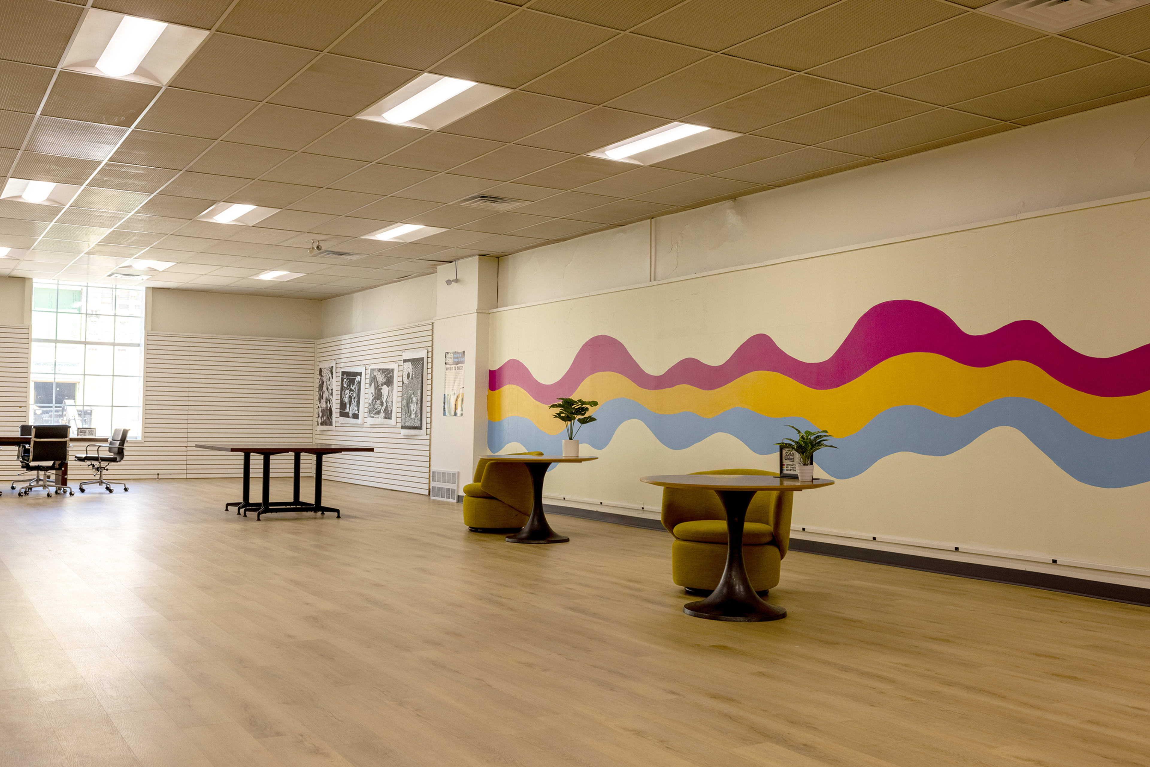 The image shows a spacious indoor area featuring two round tables with chairs, a colorful wavy mural on the wall, and large windows allowing natural light to enter.