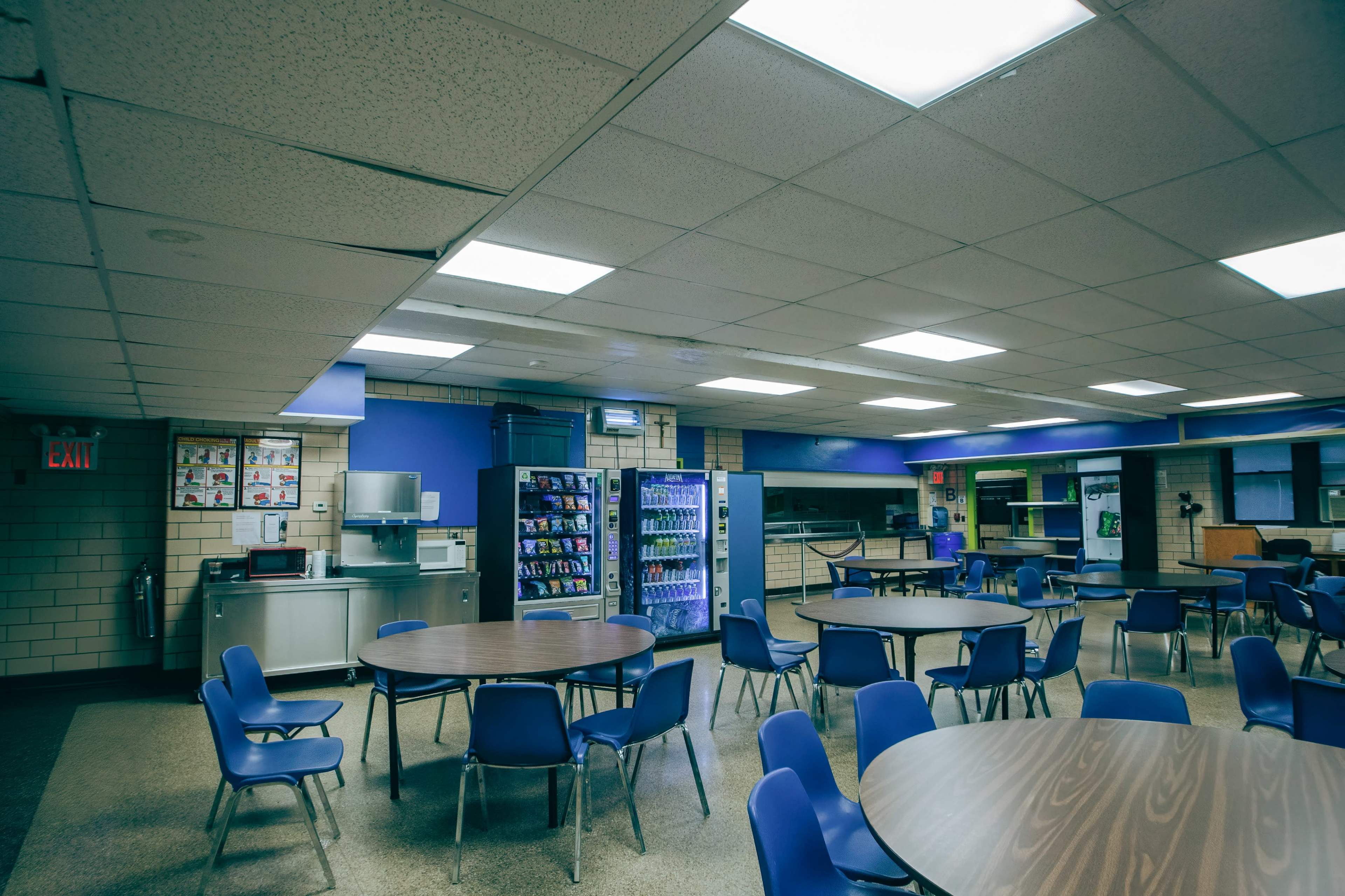 An empty cafeteria with round tables and blue chairs, a vending machine, and a kitchenette area.