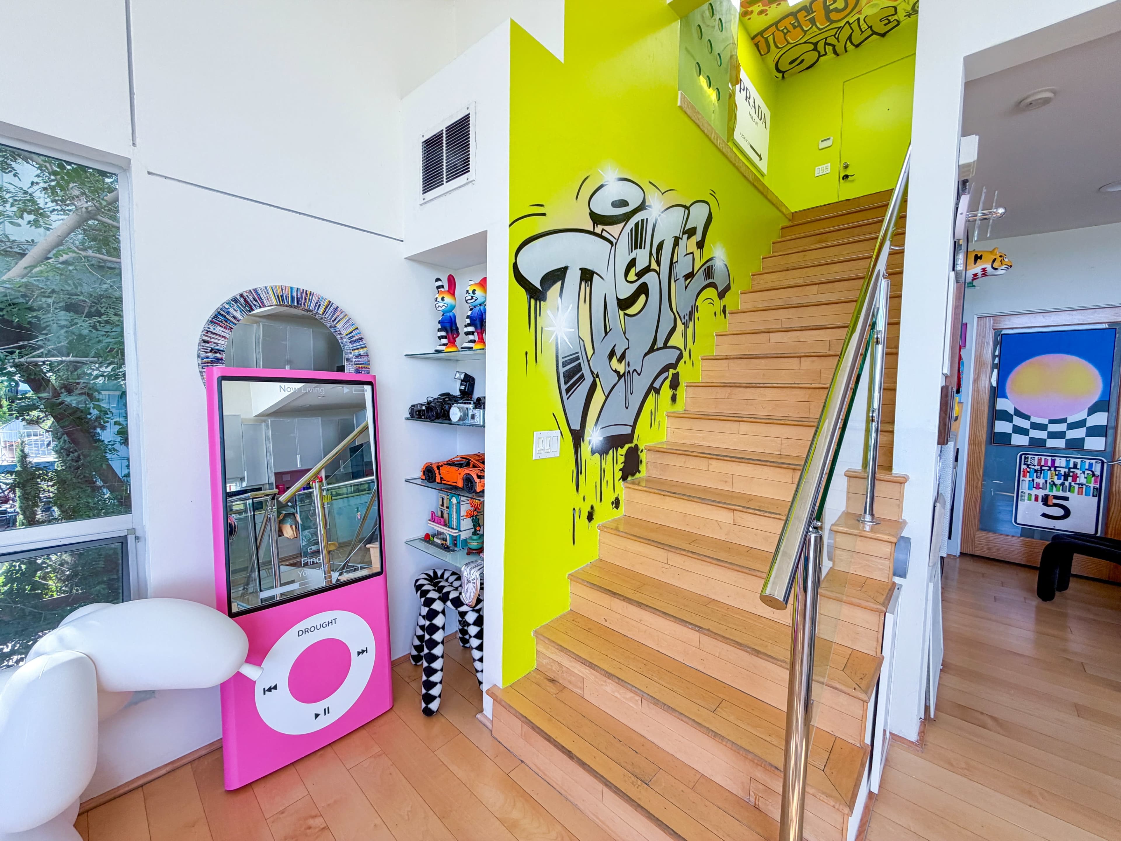 The image shows a vibrant interior space with a staircase leading to an upper level, featuring a bright green wall with graffiti, a large pink mirror, and various decorative items on display.
