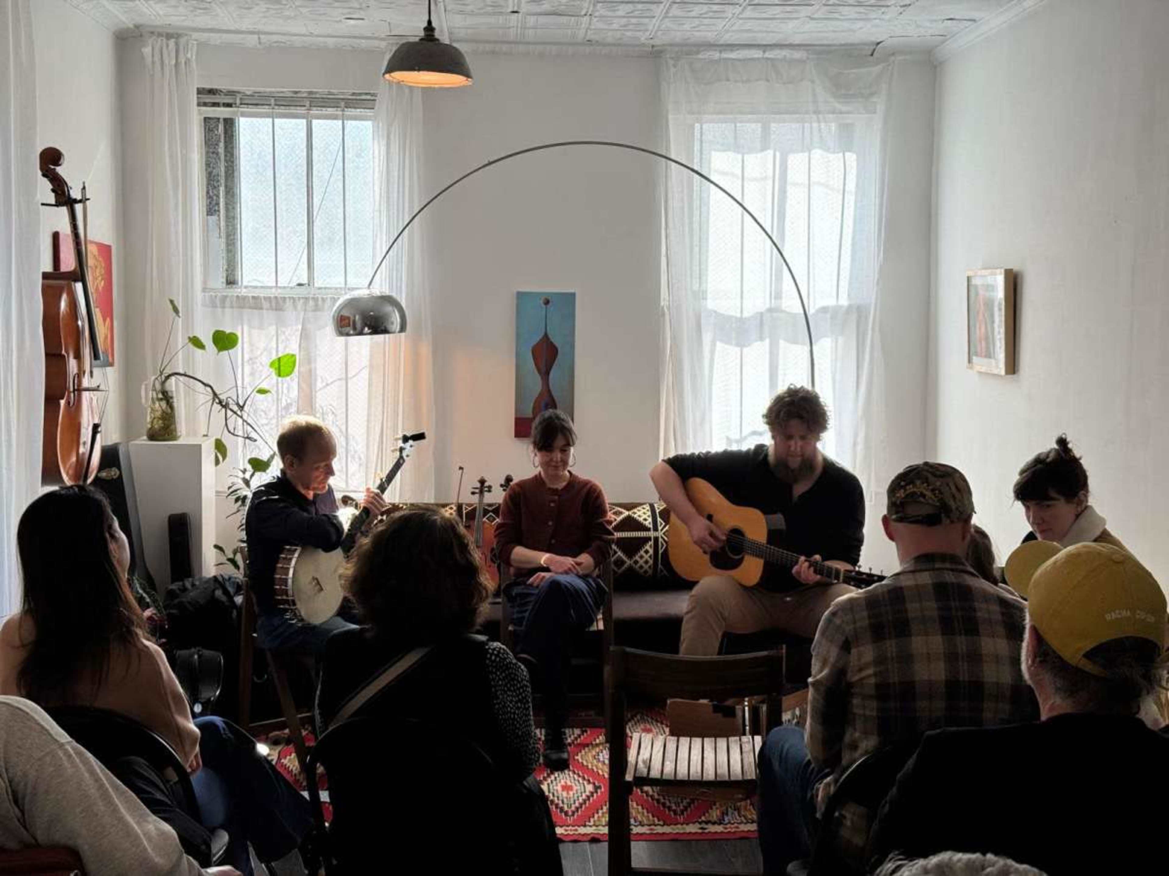 A group of musicians performs in a cozy room with a guitar, banjo, and seated audience.