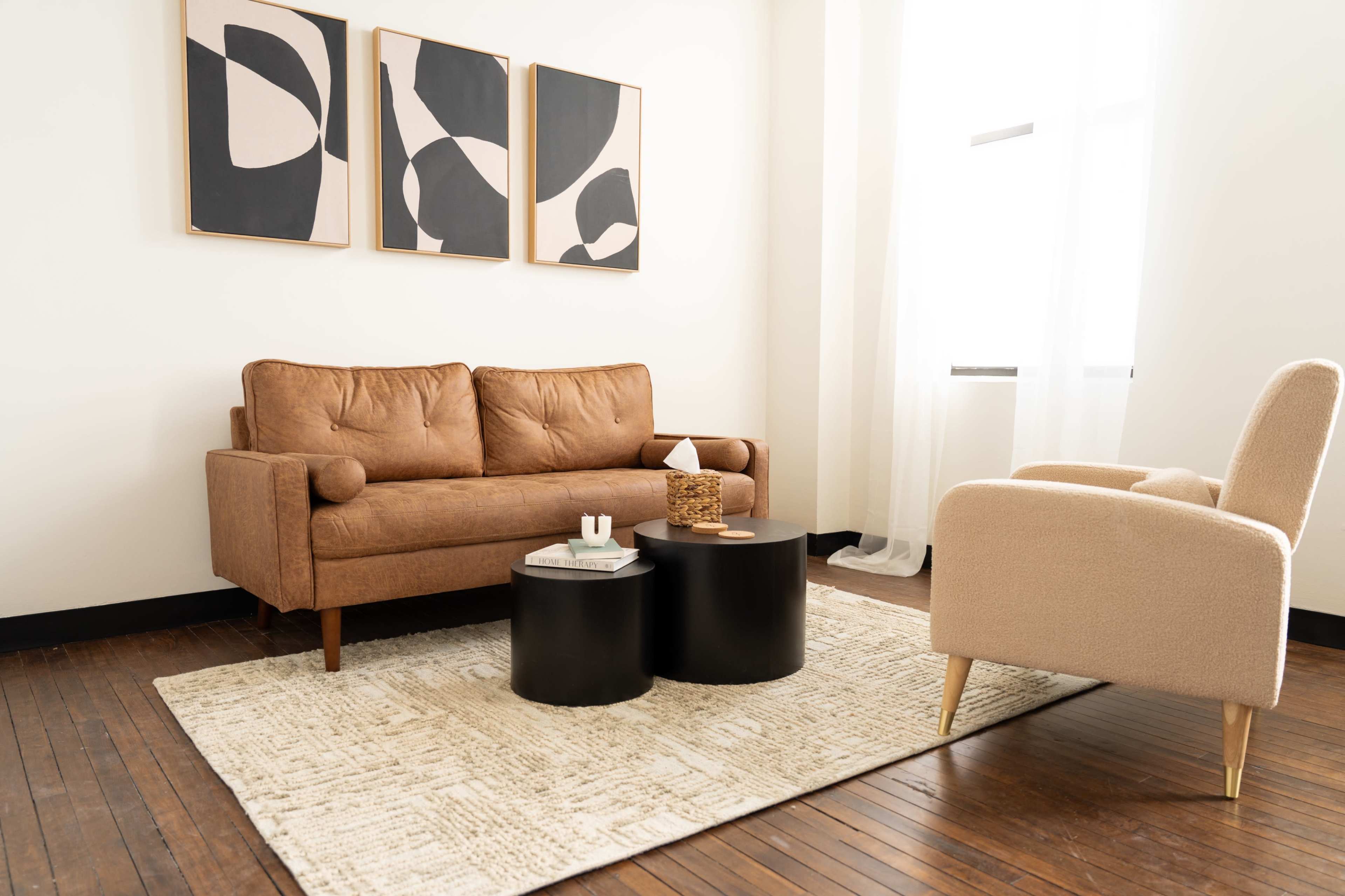 A cozy living room features a brown sofa, two black side tables, and a light-colored rug, with abstract wall art displayed above.