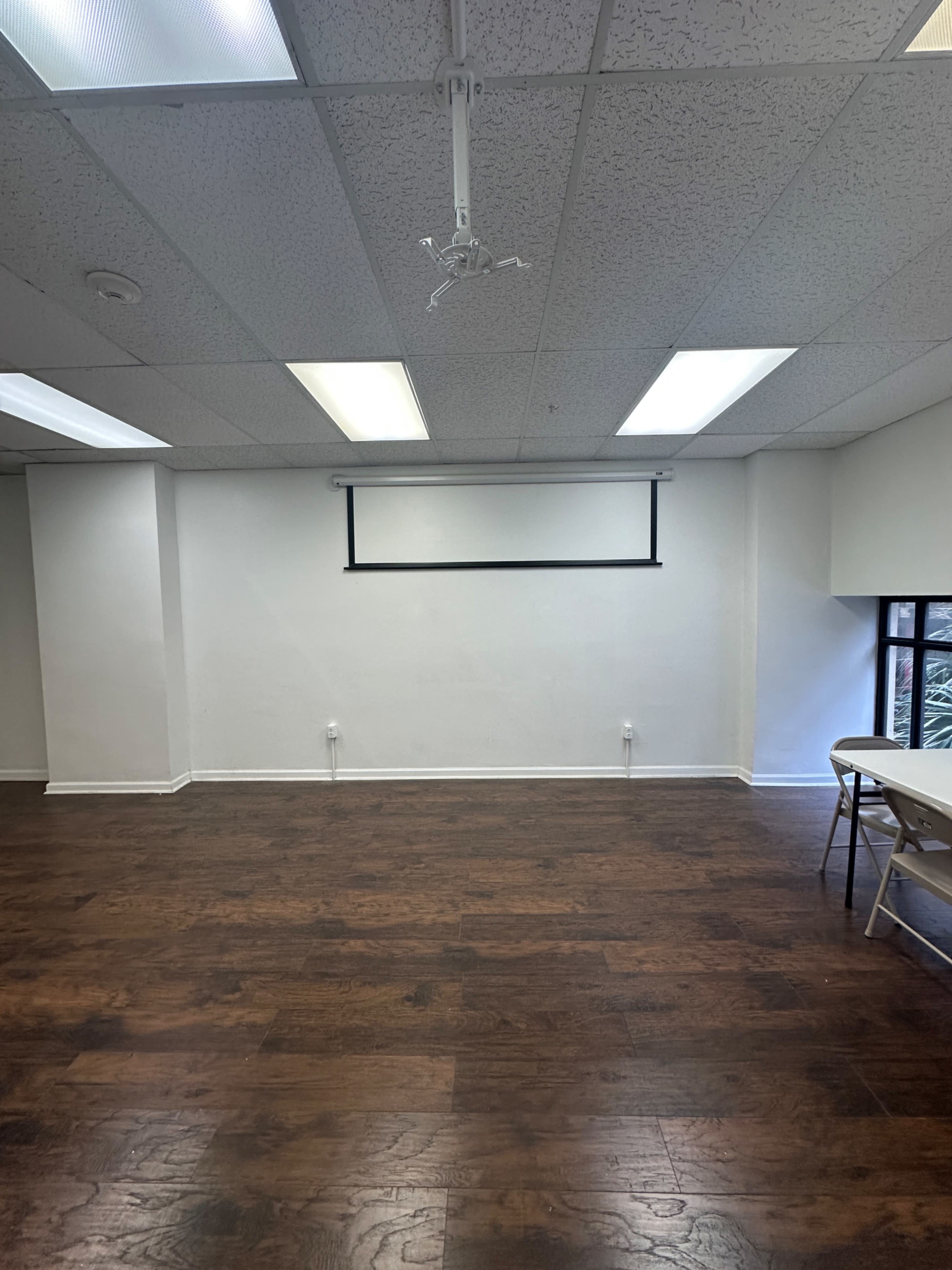 The image shows a large, empty room with wooden flooring, white walls, and a ceiling-mounted projector screen.