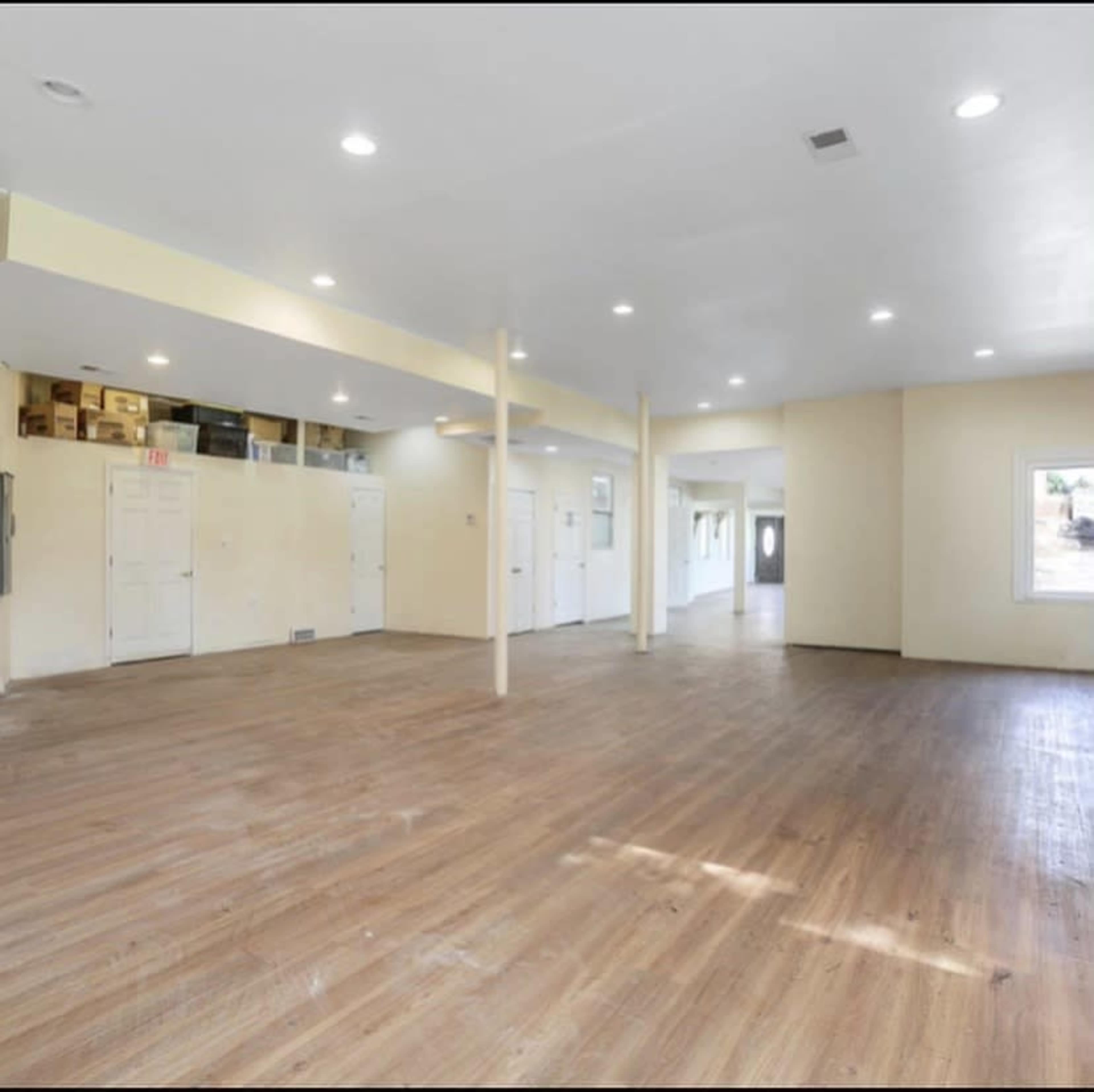 The image shows a spacious, empty room with wooden flooring, multiple doors, and bright overhead lighting.