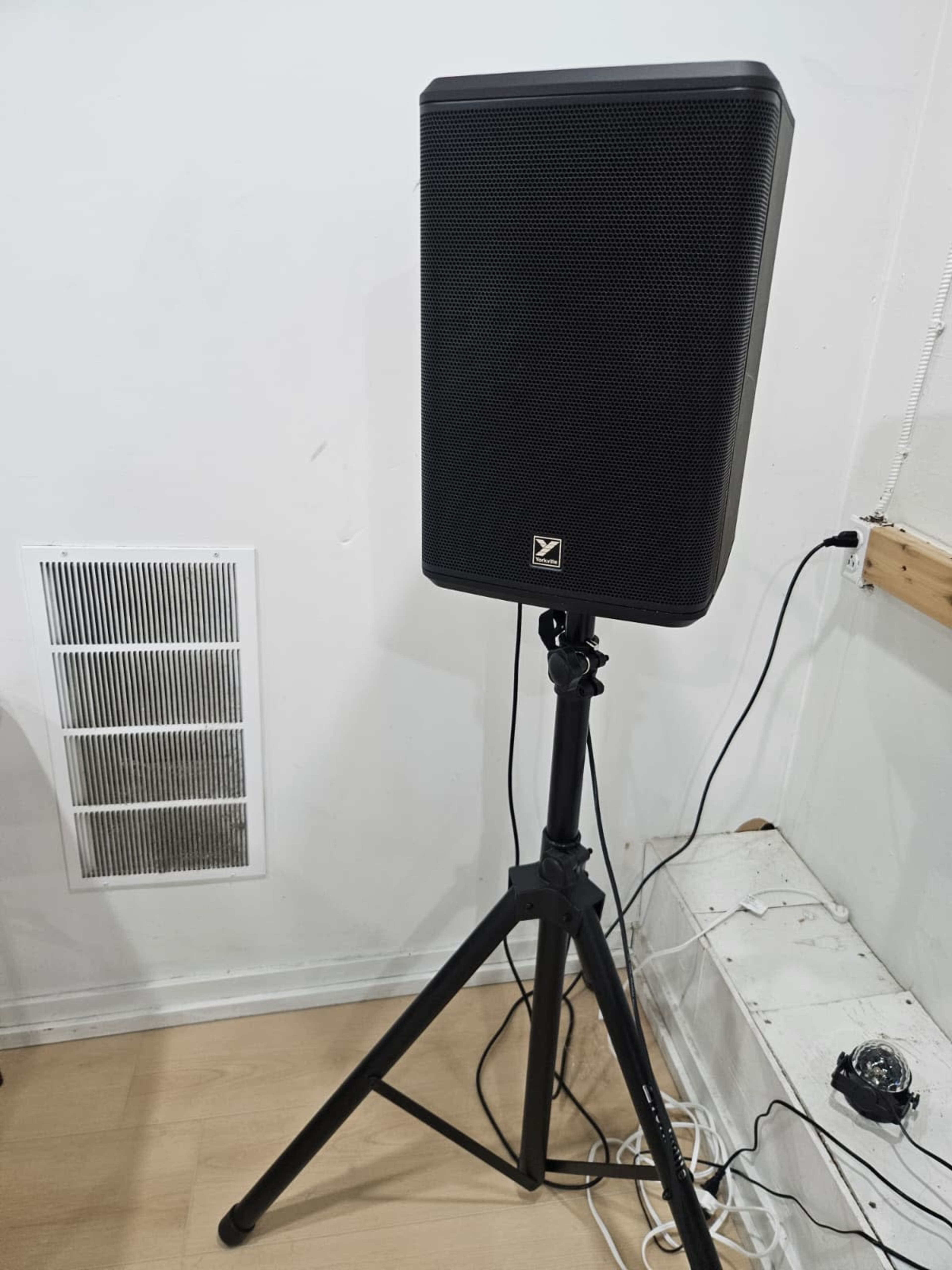 A black speaker is mounted on a black tripod stand next to a wall with a vent and electrical wires visible.