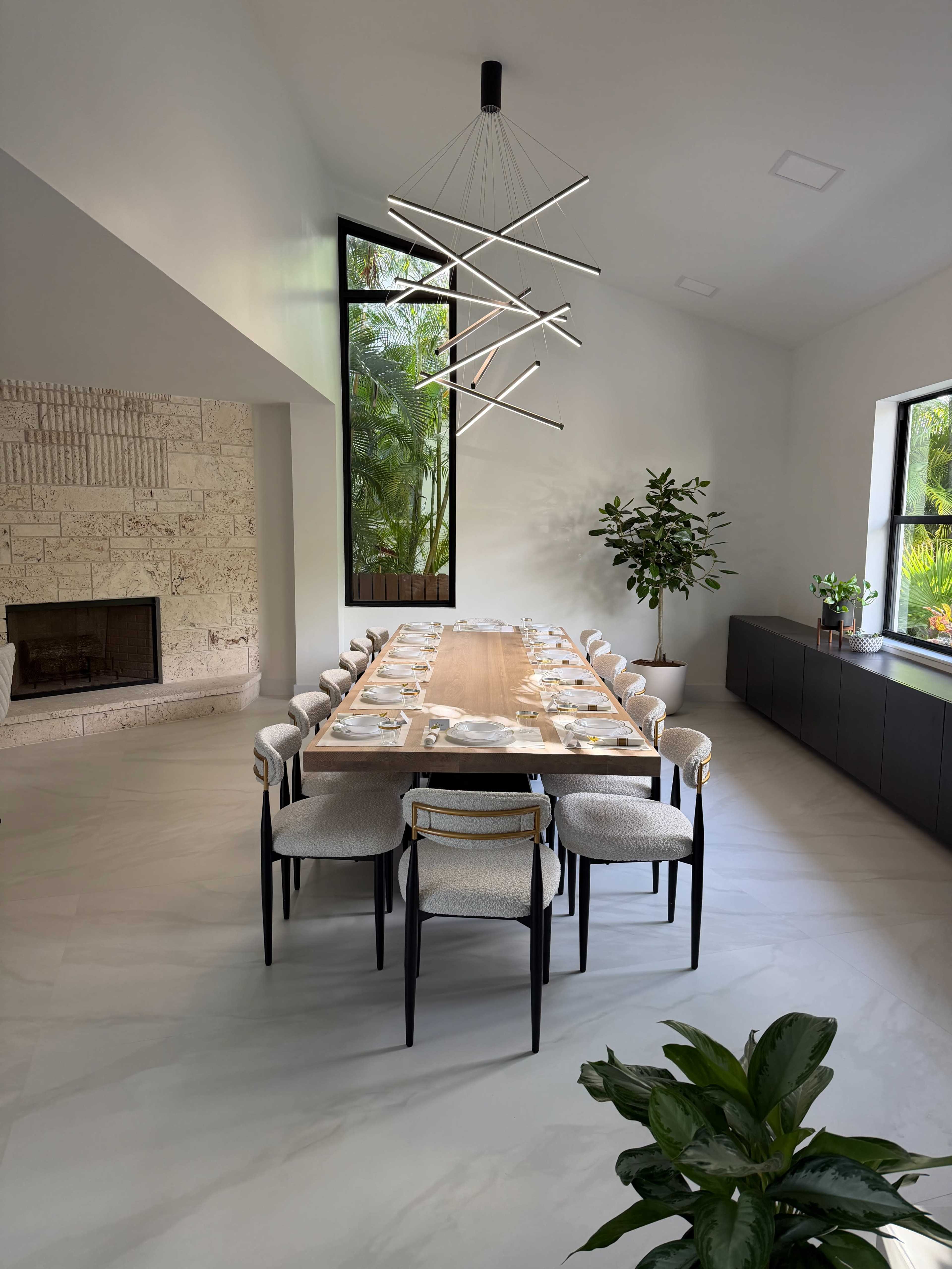 The image shows a spacious dining room with a long wooden table surrounded by contemporary chairs, a large window overlooking greenery, and modern light fixtures hanging from the ceiling.