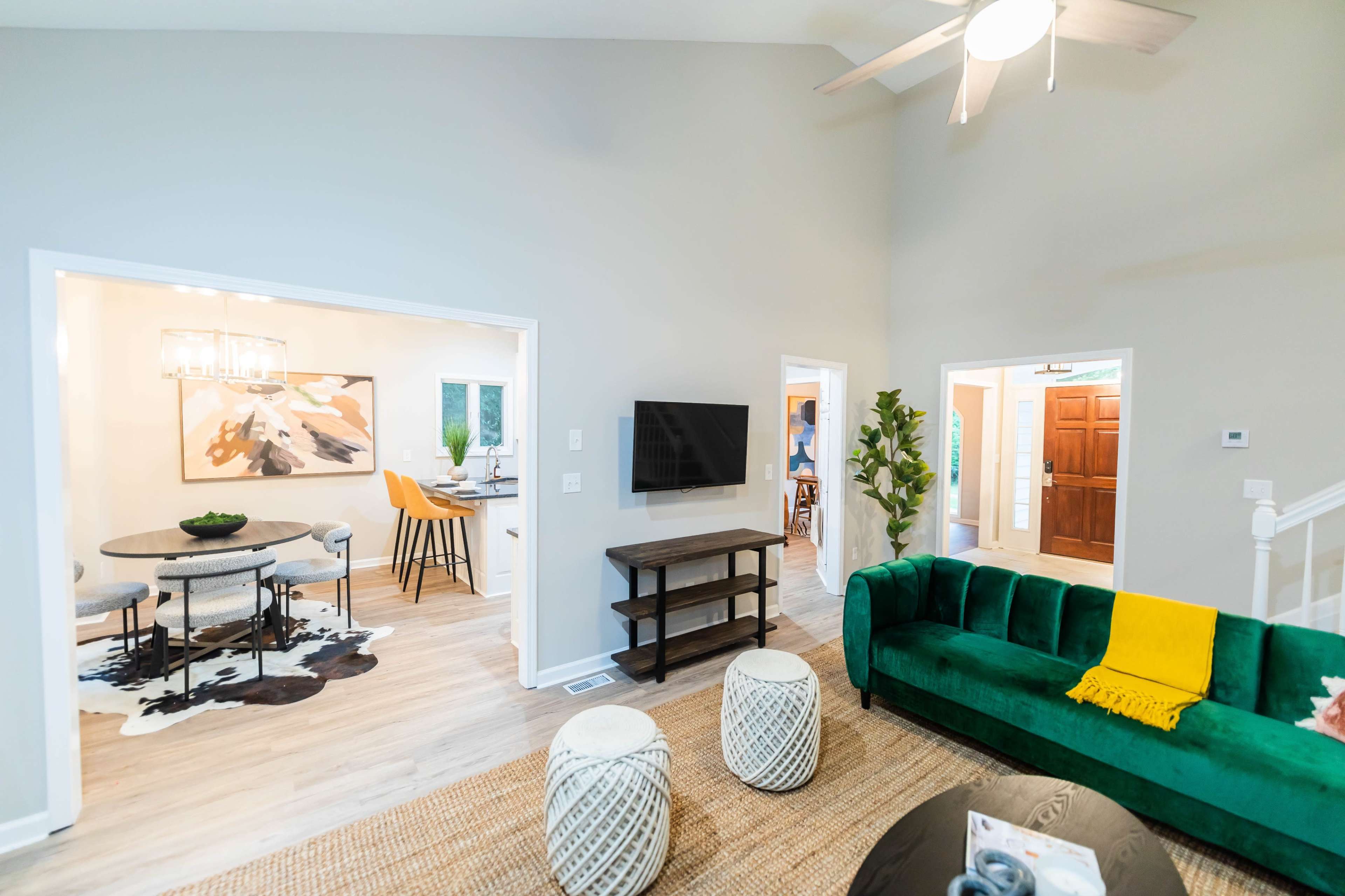 A modern living room features a green couch, a flat-screen TV on a wooden stand, and woven poufs, with open access to adjacent dining and entry areas.