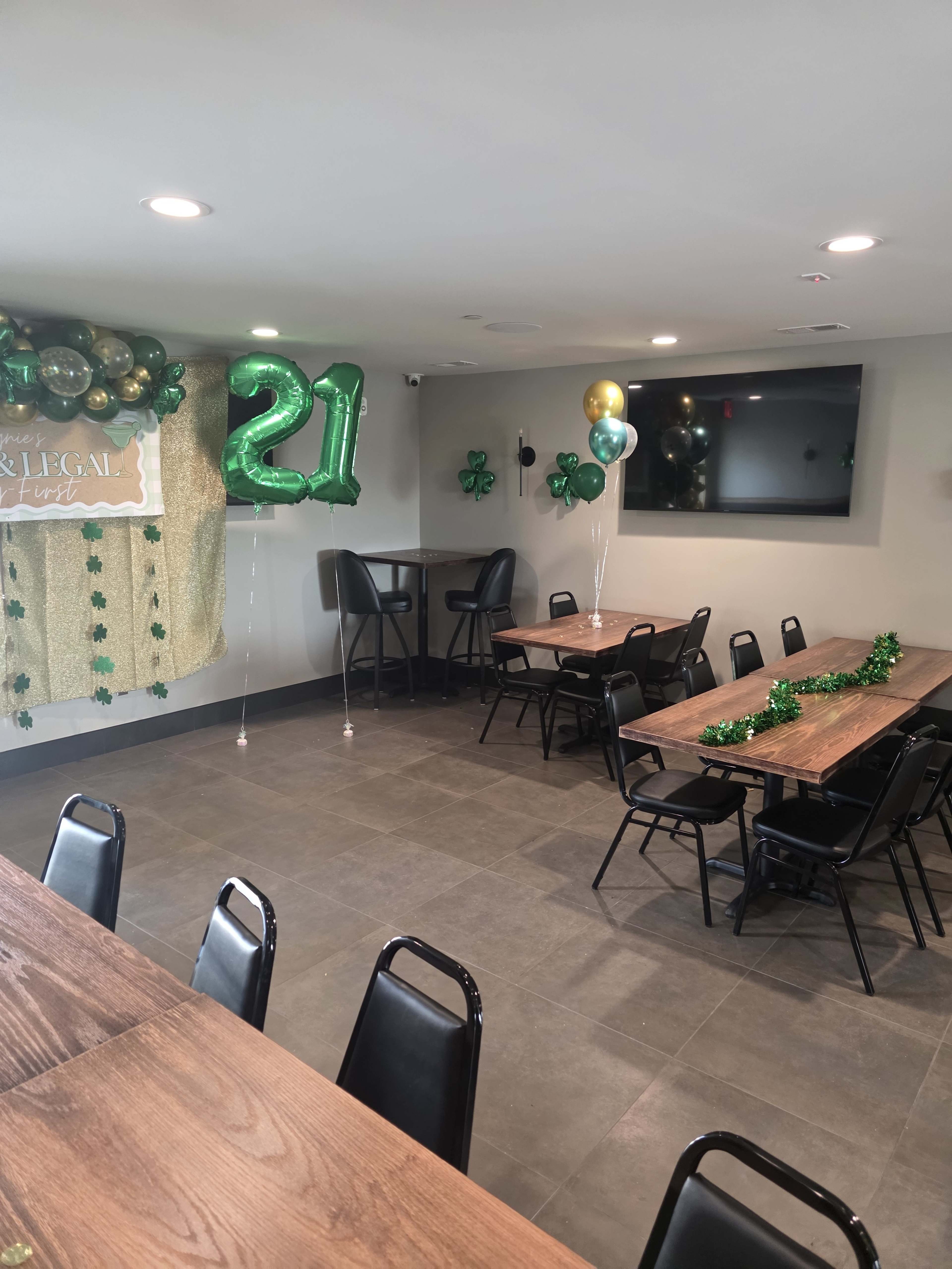 The image shows a decorated party room featuring tables and chairs, with green balloons, shamrocks, and a large "21" balloon.