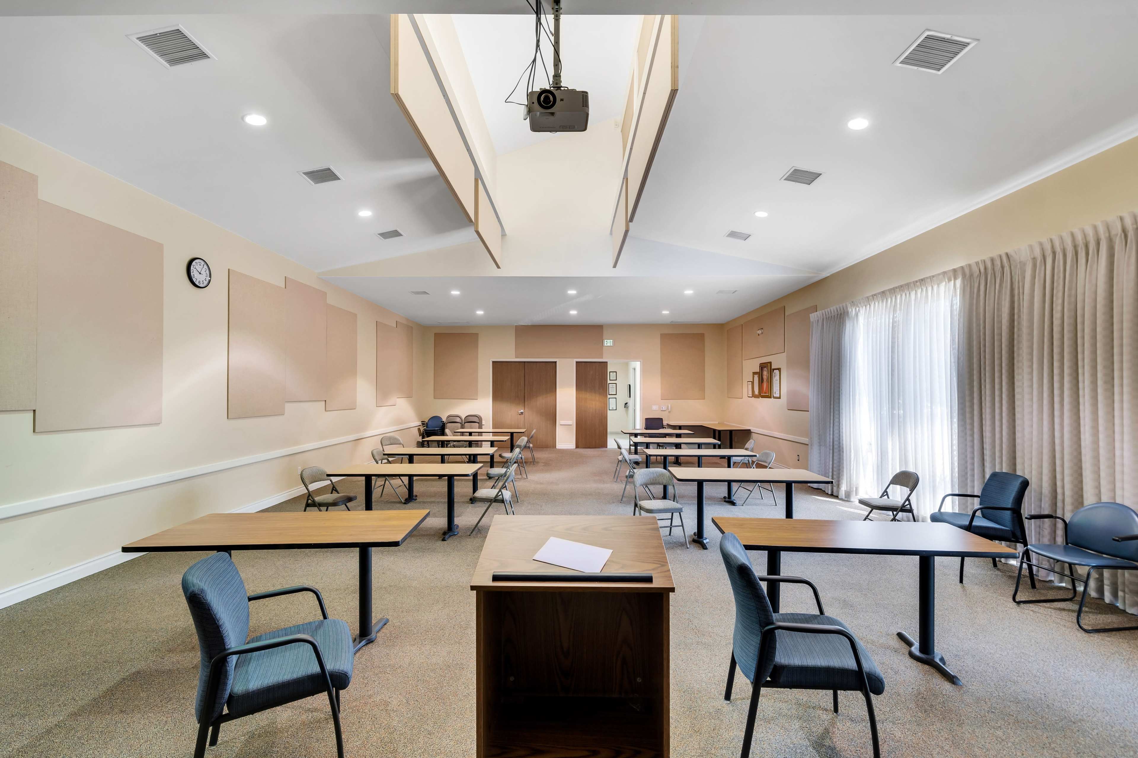 A spacious meeting room with several tables and chairs arranged for a gathering, featuring a clock on the wall and large windows with curtains.