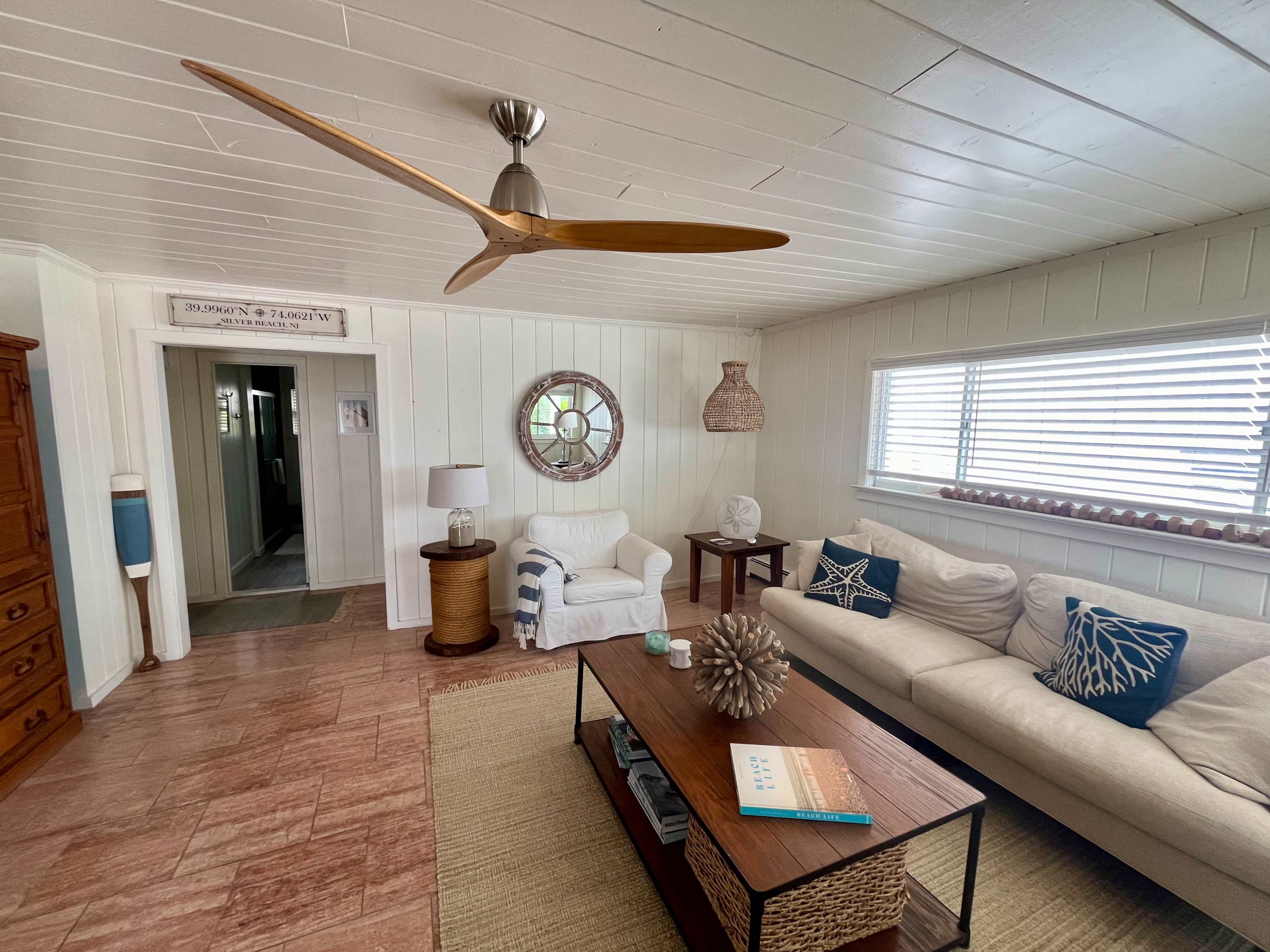 A light-colored living room with a ceiling fan, a coffee table, a couch, and decorative elements including pillows and a mirror.
