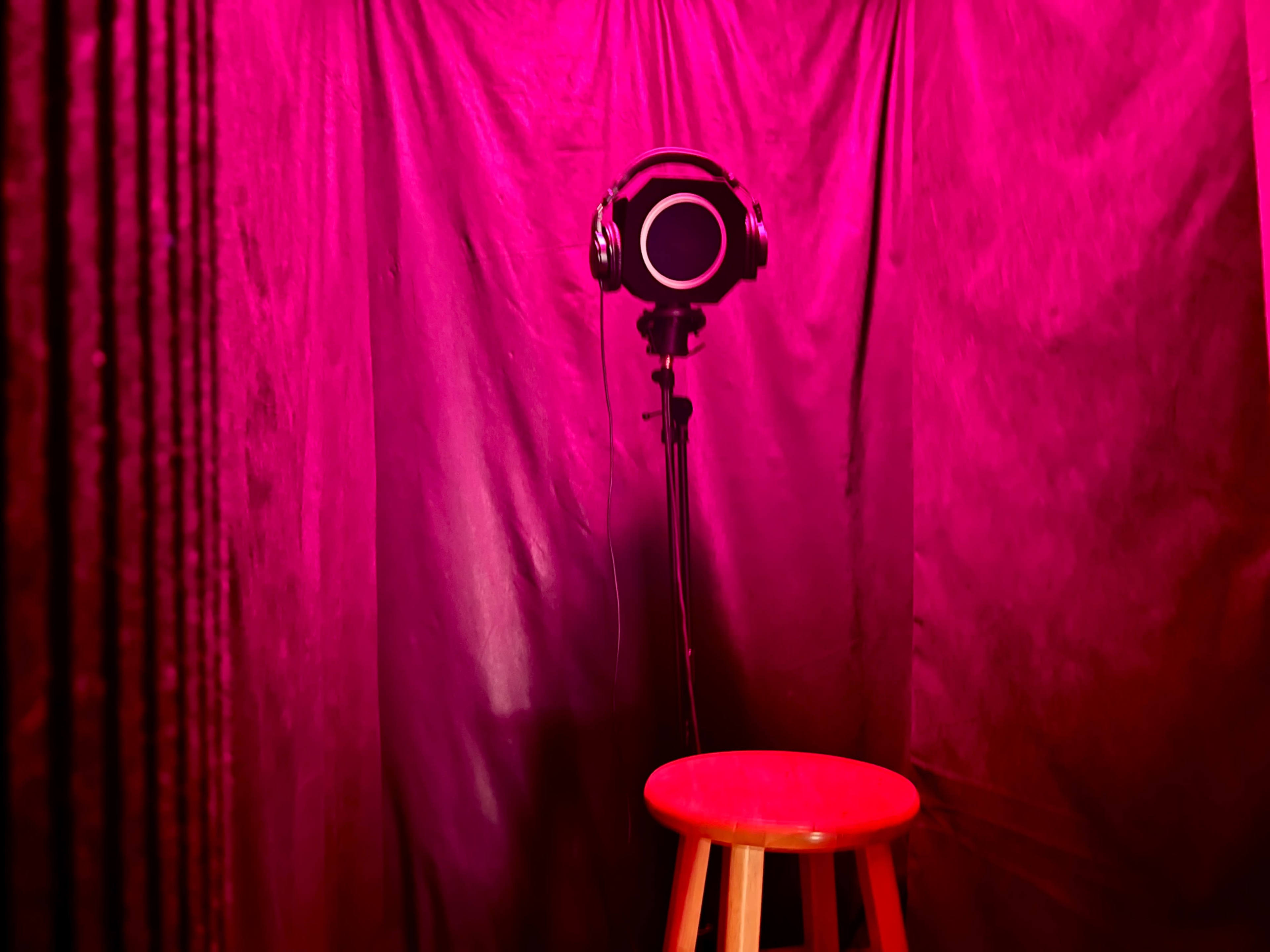 A black microphone with headphones is mounted on a stand in front of a red-lit curtain, above a wooden stool.