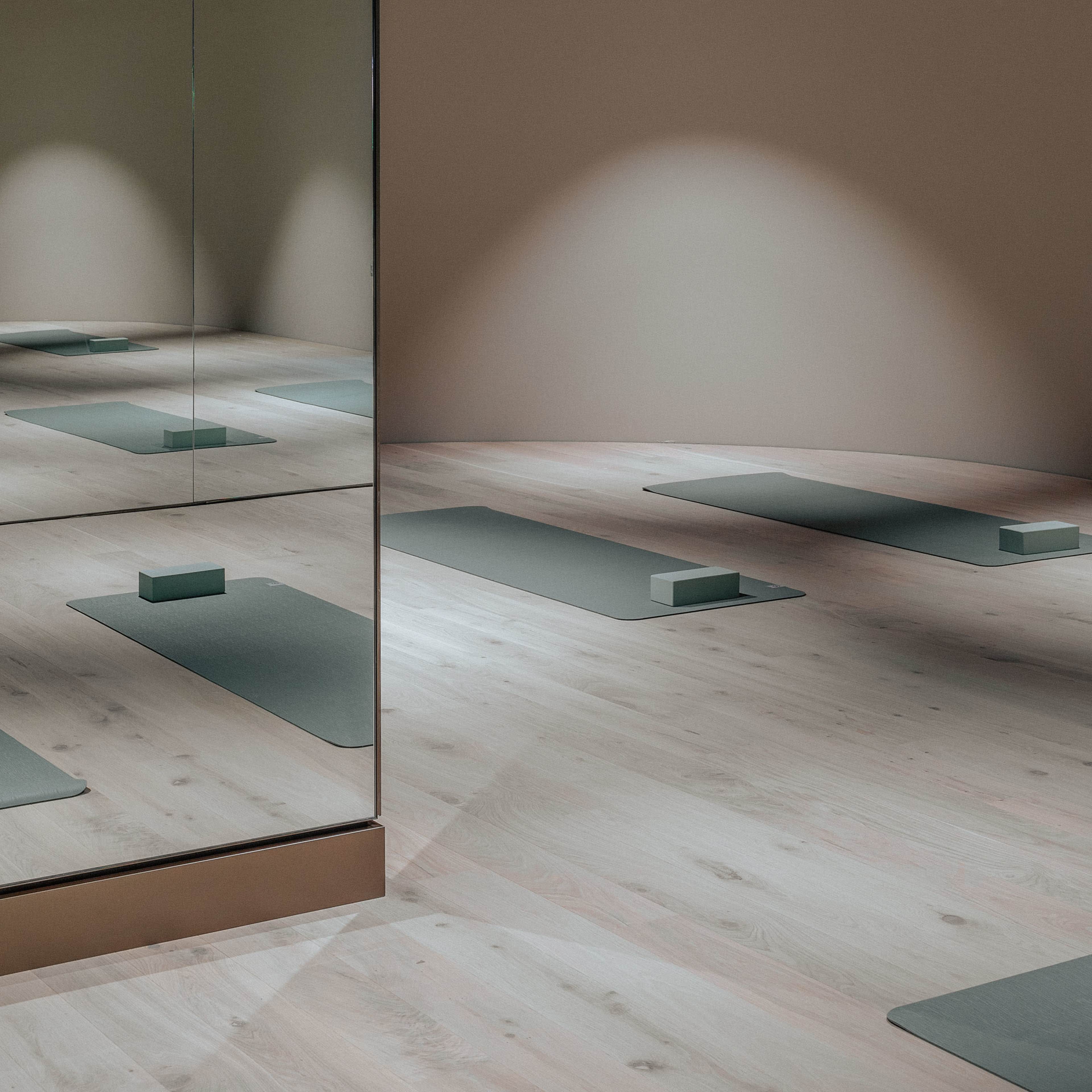 The image shows a sparsely furnished yoga studio with several yoga mats aligned on a light wooden floor and a mirror on one wall.