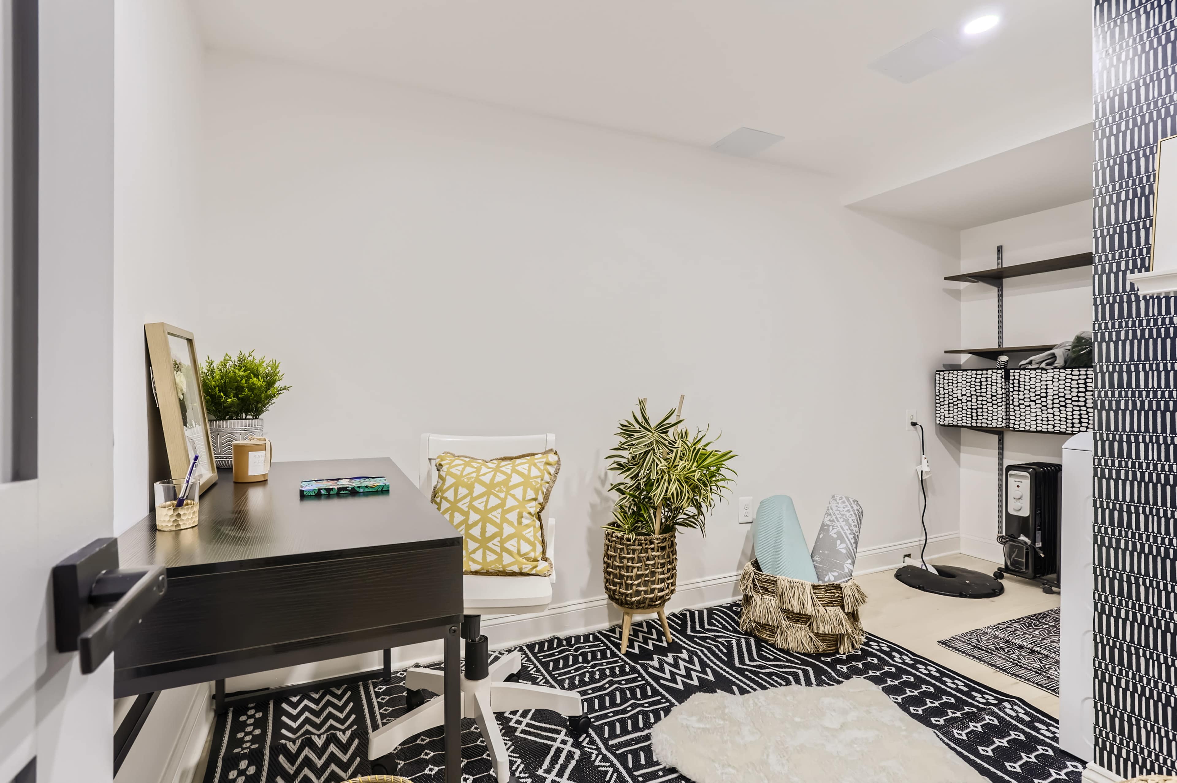 A minimalist home office setup featuring a desk, a chair, decorative plants, and patterned rugs against a neutral wall.