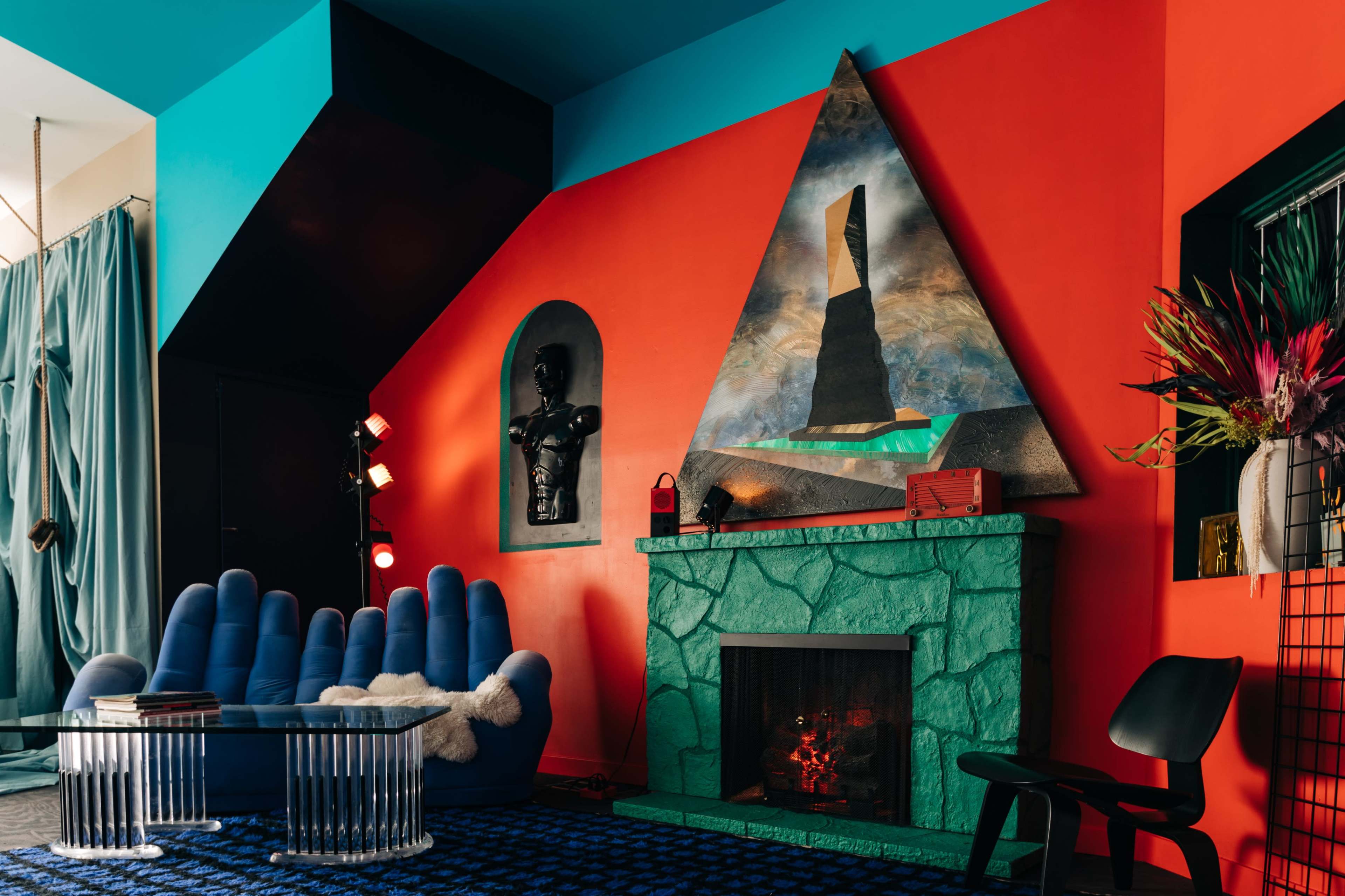 The room features a vibrant orange wall with a triangular artwork above a green stone fireplace, complemented by a blue sectional sofa and a glass coffee table.