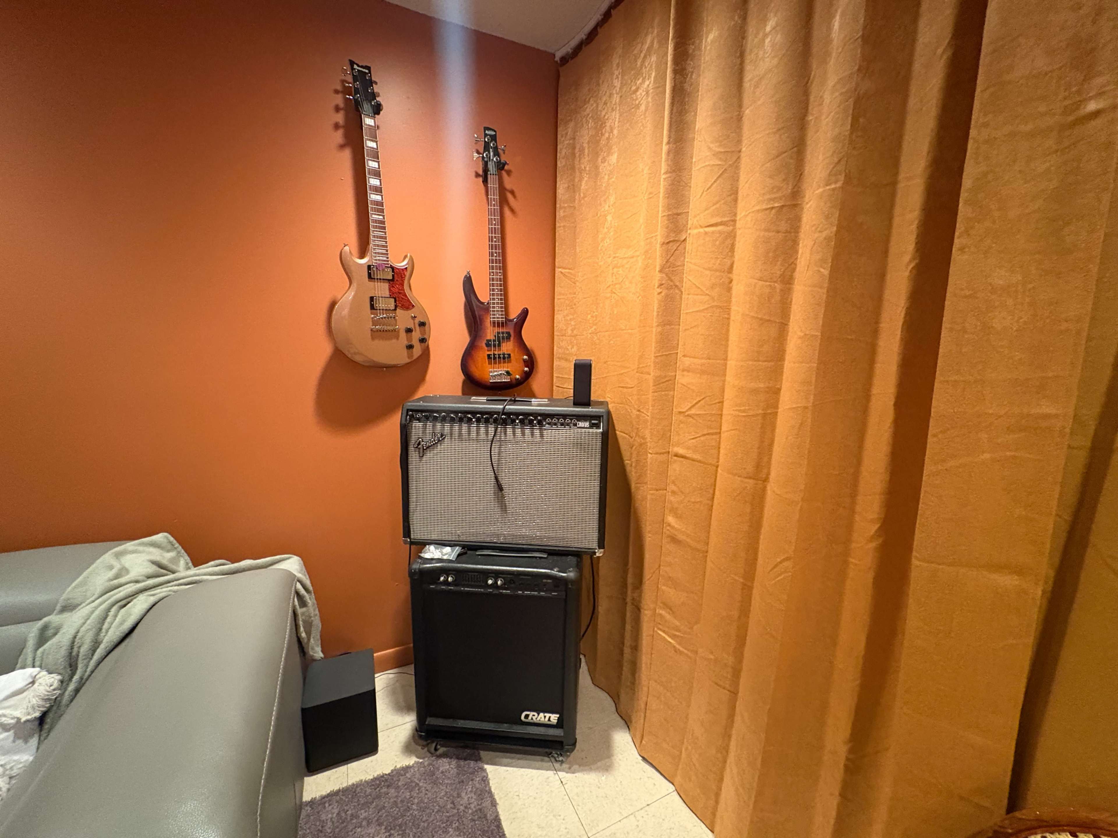 The image shows two electric guitars hanging on a wall beside an amplifier and a speaker, with a couch and a curtain in the corner of a room.
