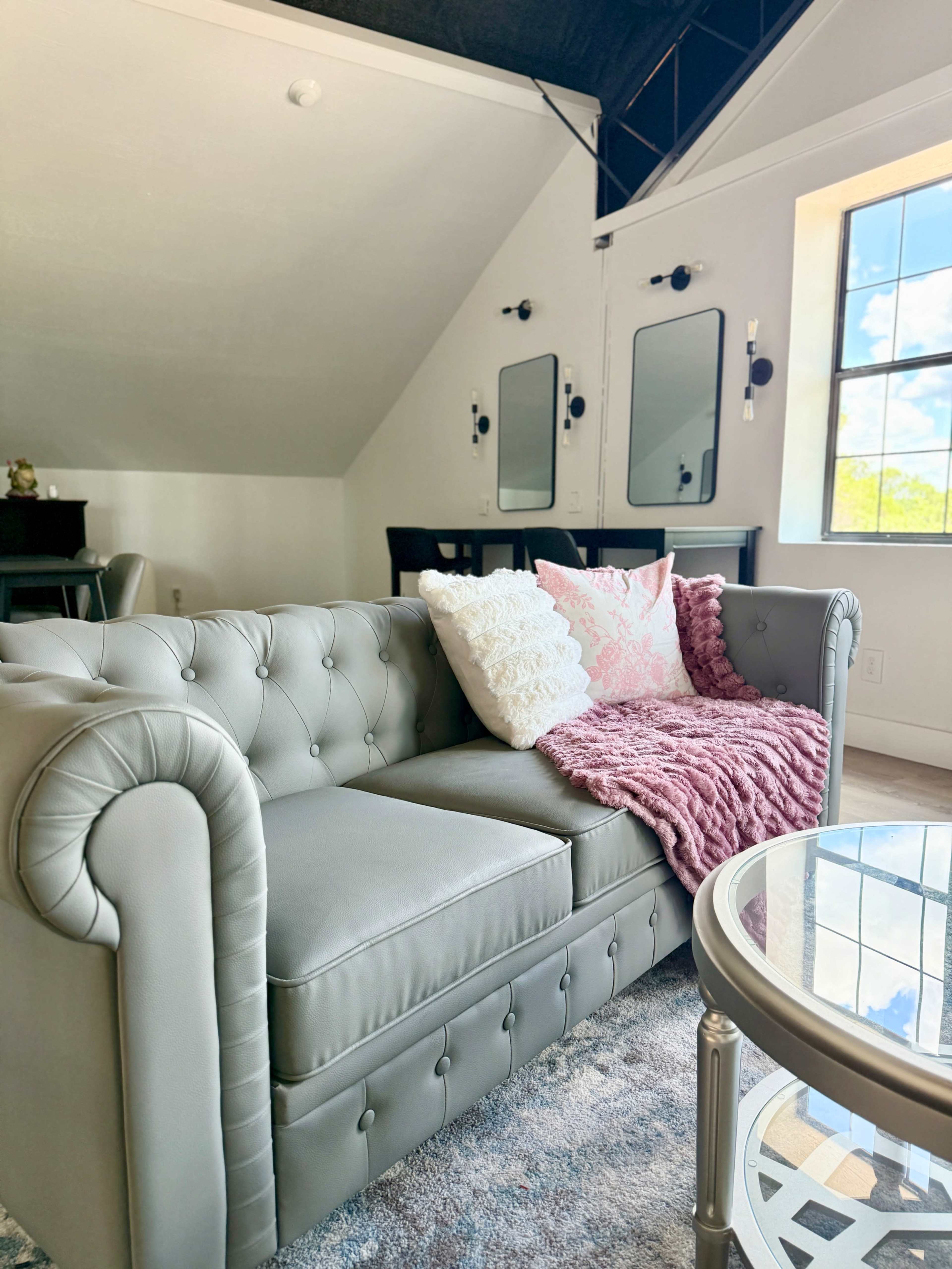 The image shows a modern living room featuring a gray tufted sofa with decorative pillows and a pink throw, accompanied by a round glass coffee table and large windows allowing natural light.