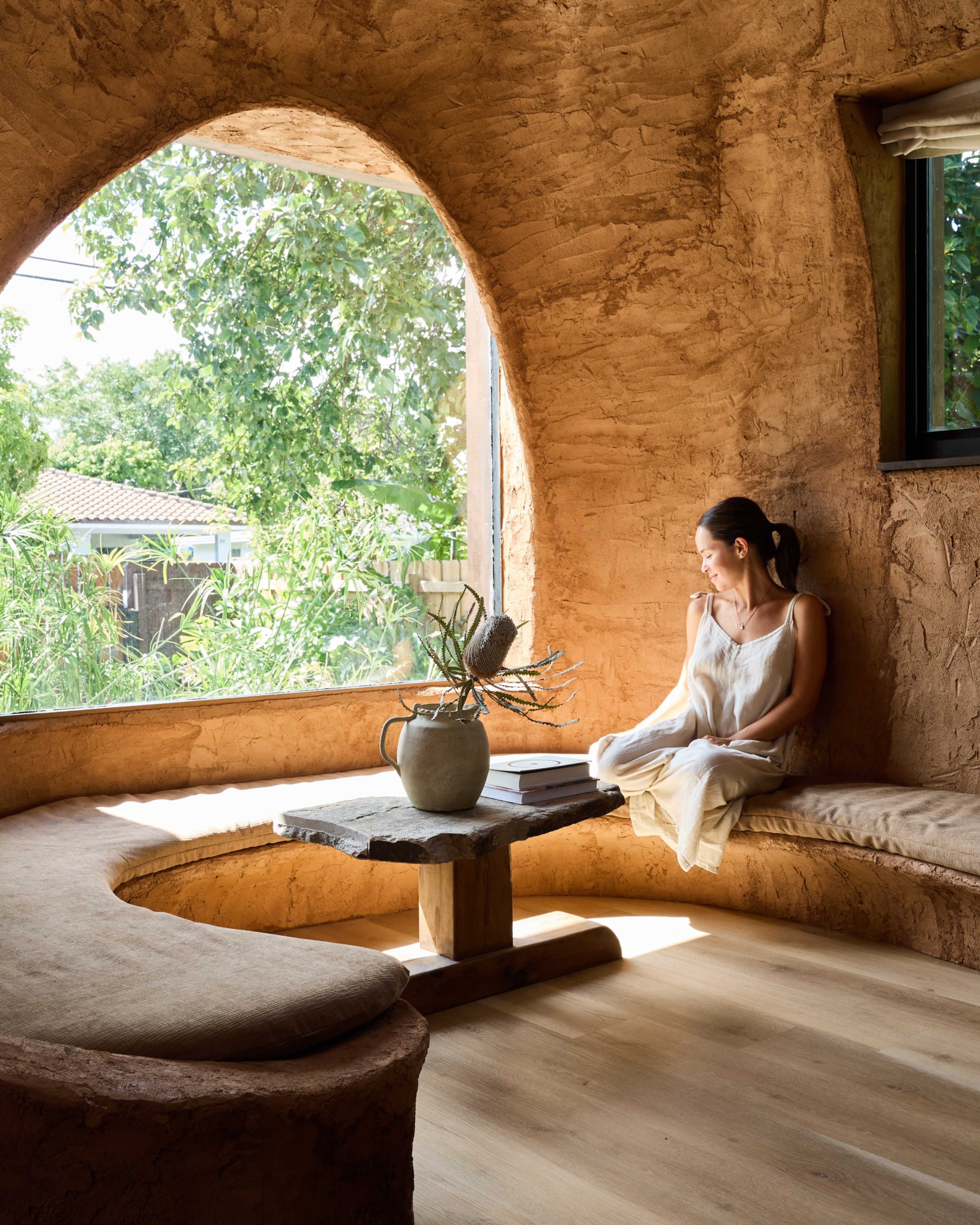 A woman seated on a curved bench beside a large window, surrounded by earthy textured walls and greenery outside.