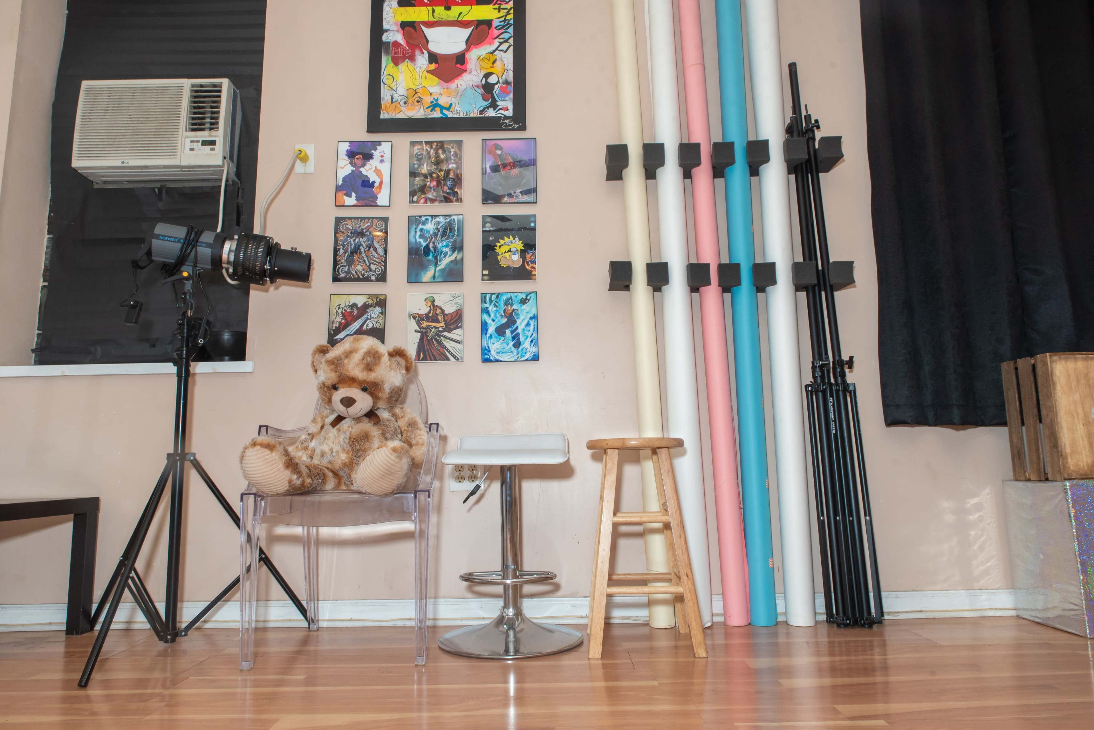 The image shows a cozy studio space featuring a clear chair with a teddy bear, a metal stool, colorful backdrop rolls, a camera on a tripod, and artwork on the wall.