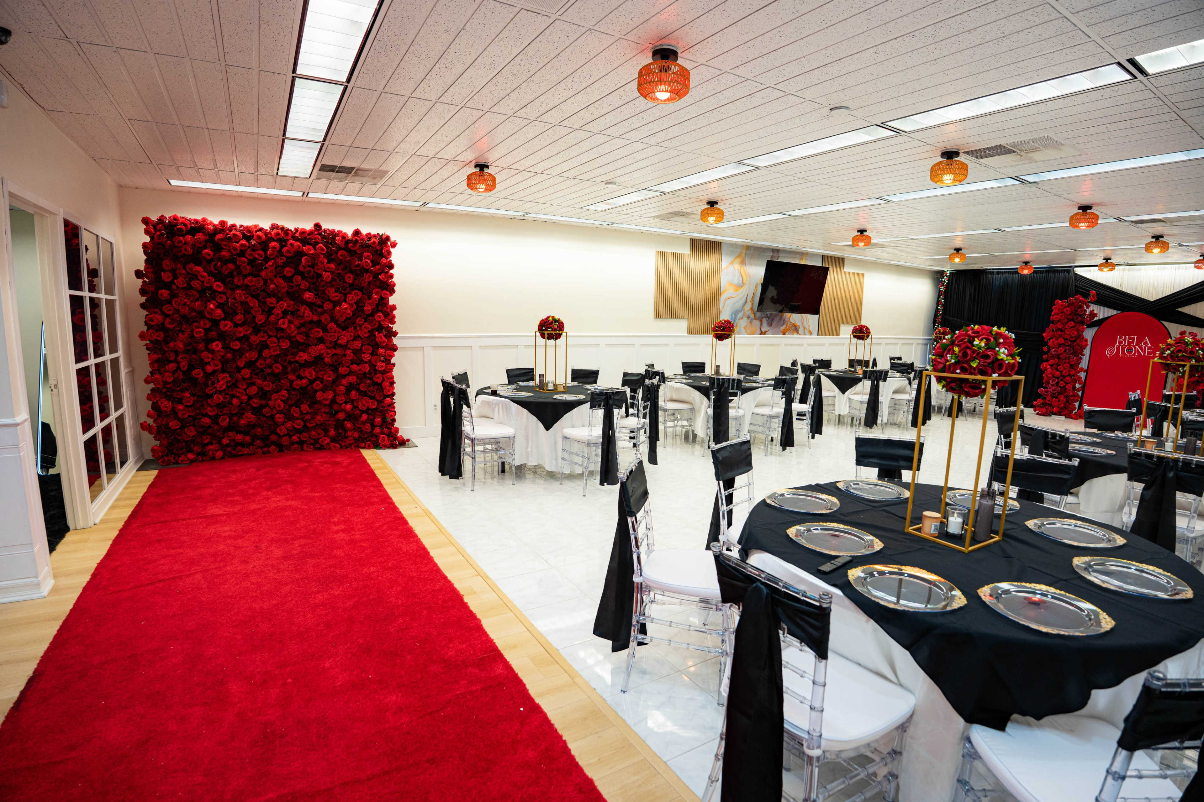 The image shows a decorated event space featuring a red carpet, tables set with black tablecloths and silver plates, and a wall adorned with artificial red roses.
