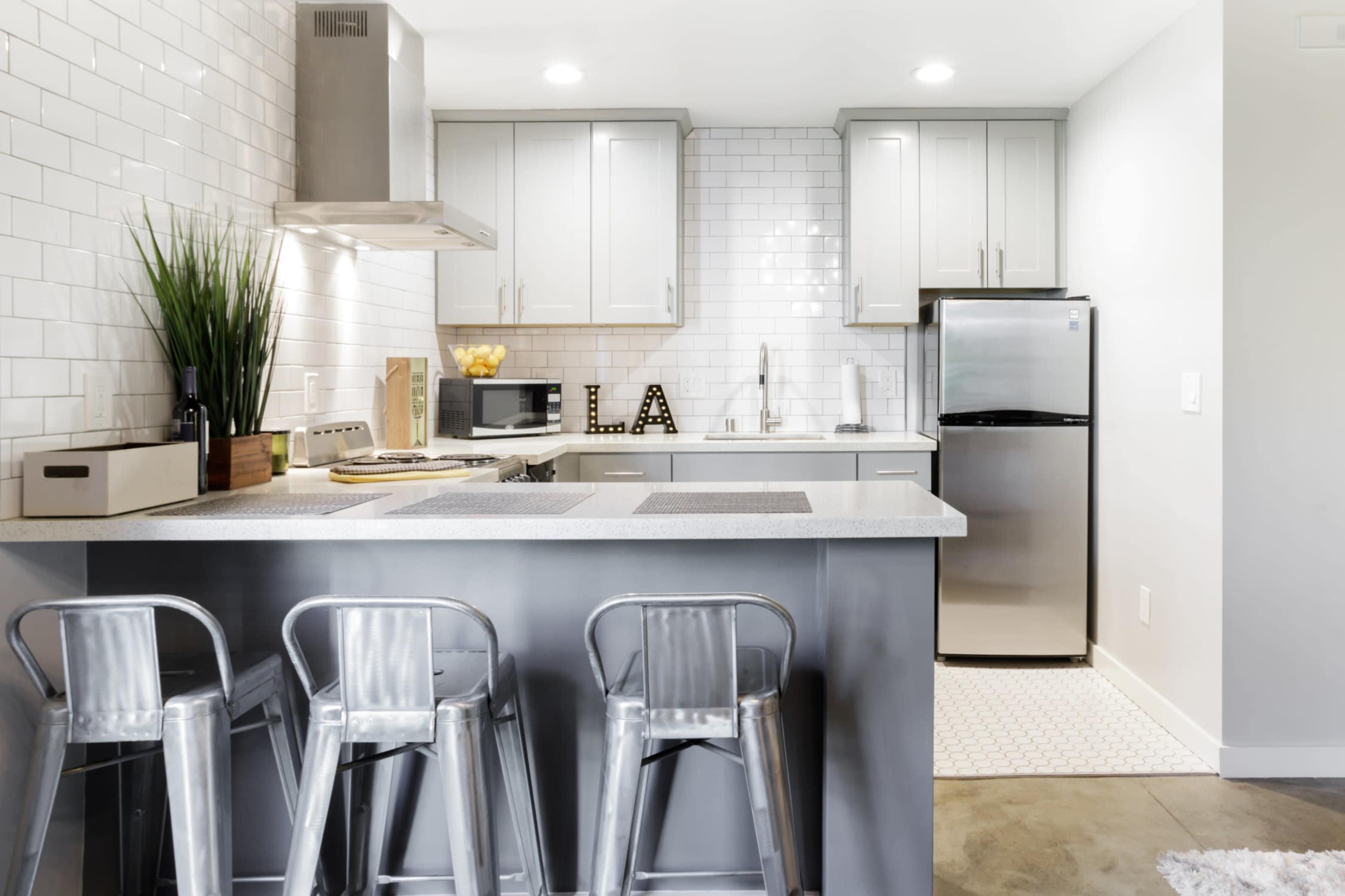 A modern kitchen features stainless steel appliances, white subway tile backsplash, and two metal bar stools at a gray island countertop.