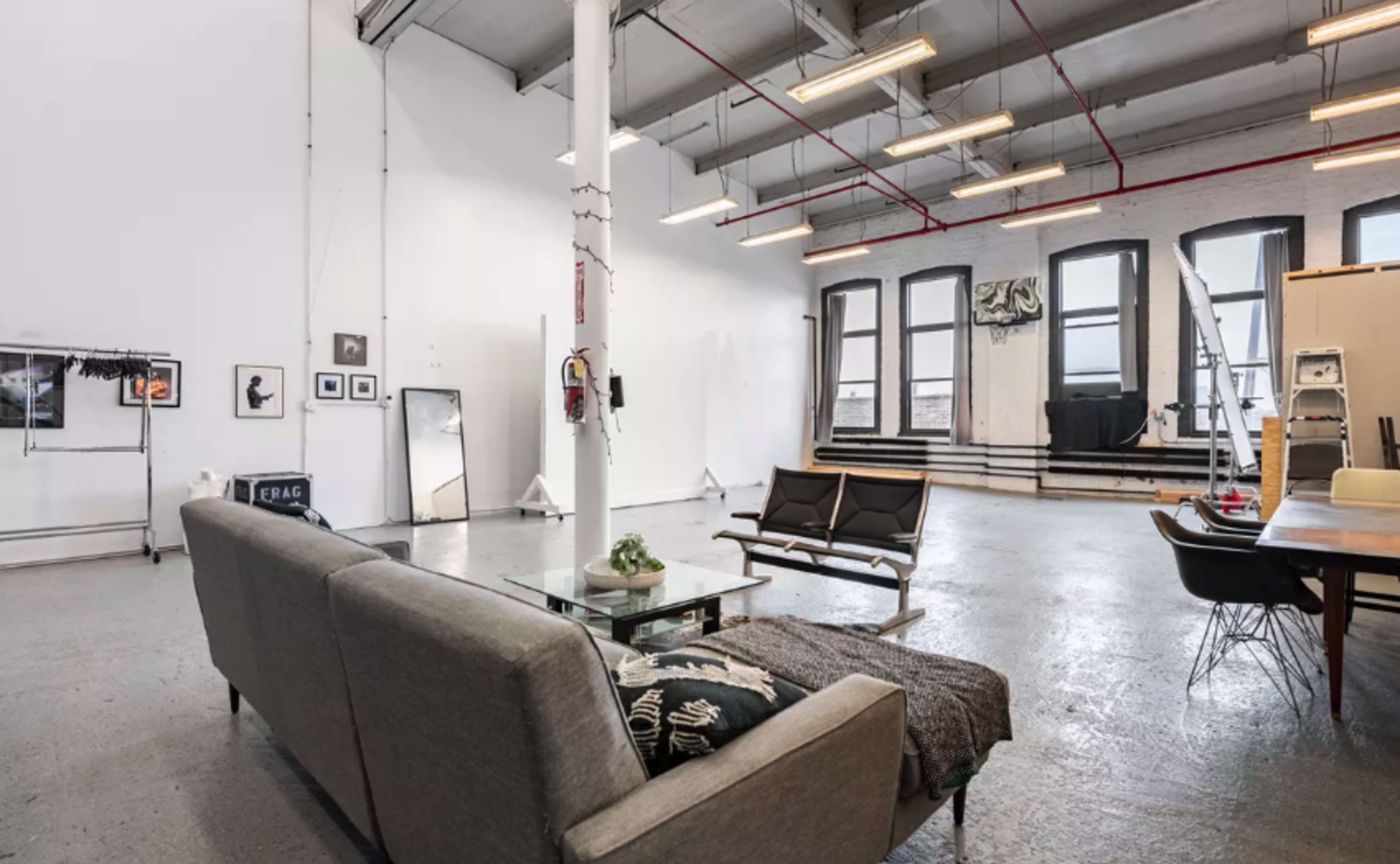 A spacious, modern studio with a gray sofa, a glass coffee table, and large windows, featuring minimalist decor and various items scattered throughout.