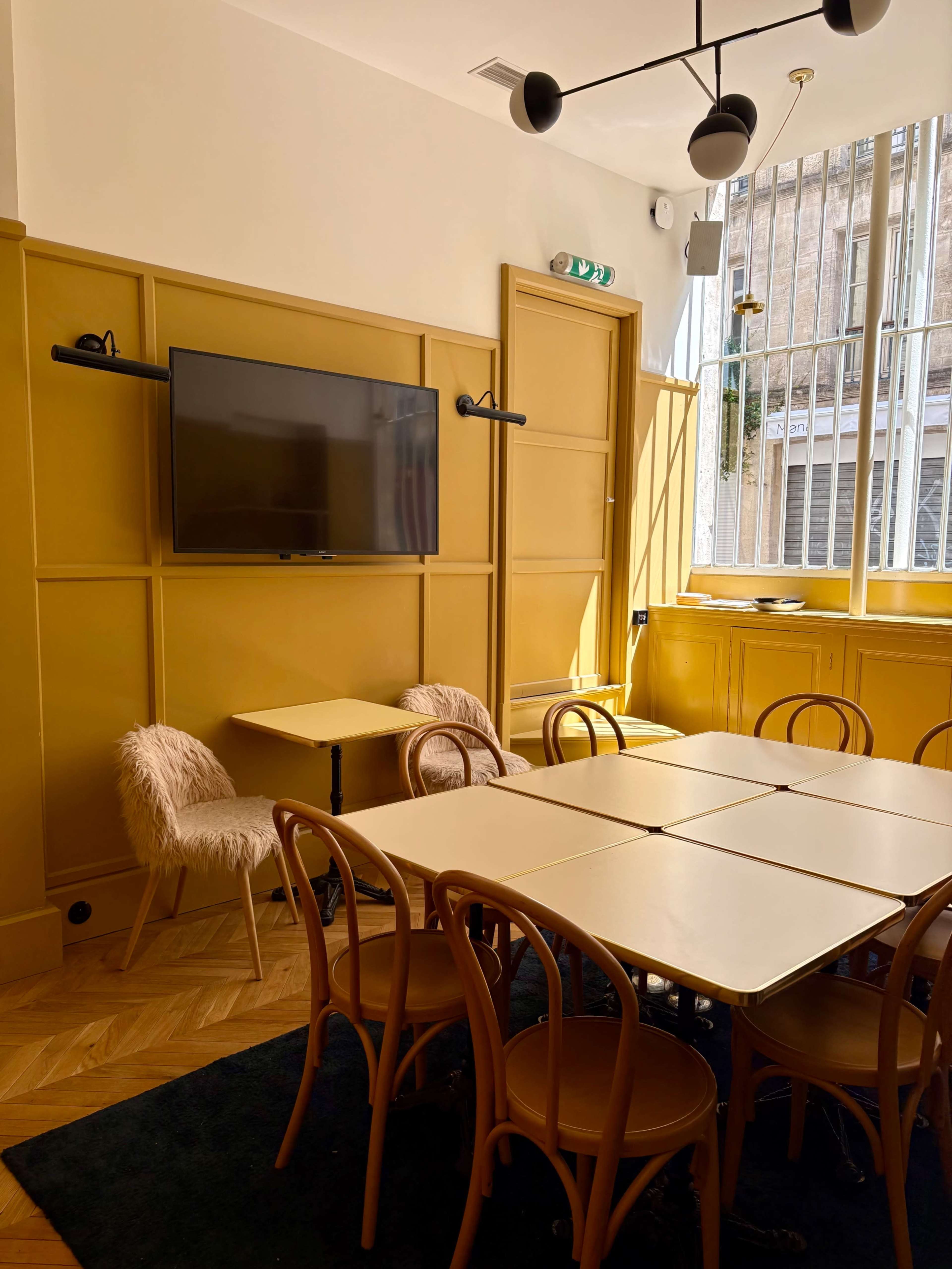 The image features a brightly lit room with yellow-paneled walls, a large TV mounted on one wall, and several tables surrounded by wooden chairs.