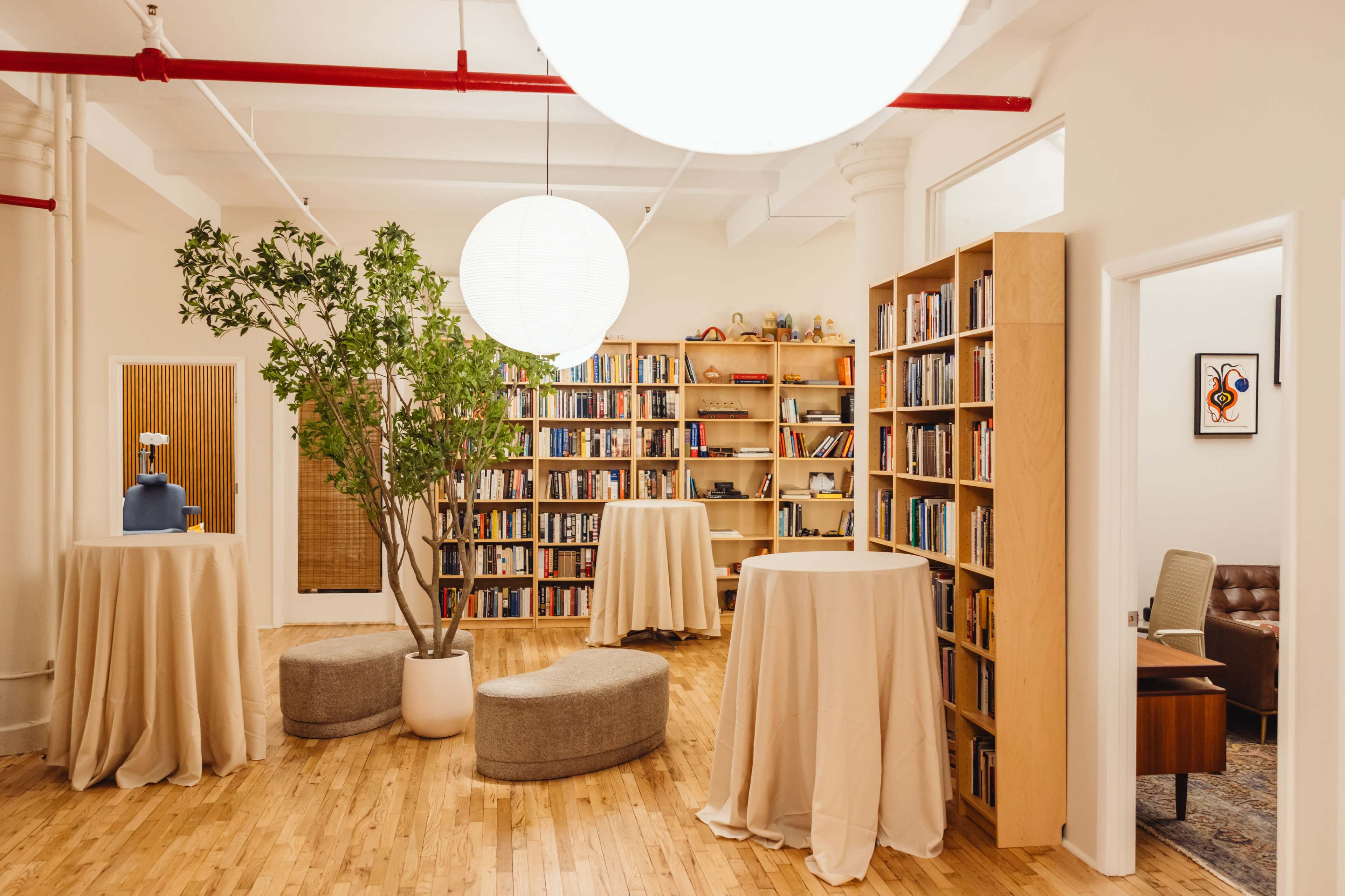 The image shows a spacious room with bookshelves filled with books, several round tables covered with beige cloths, and a central tree arrangement.